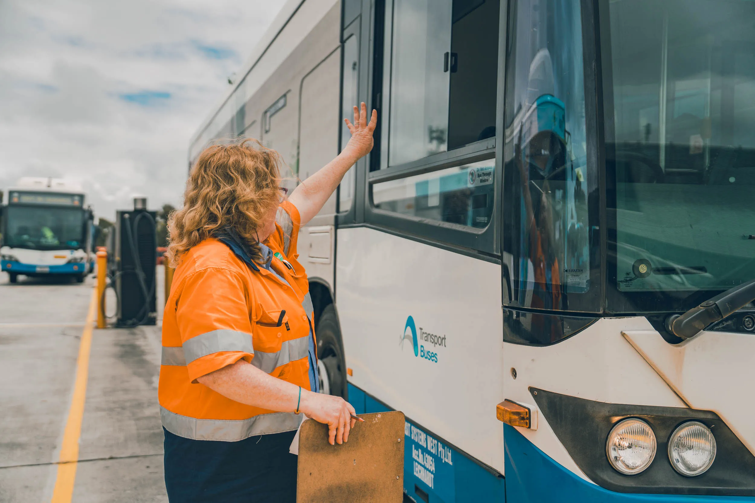 Woman in orange reflective jacket holding clipboard and raising hand to white and blue Transport Buses vehicle.