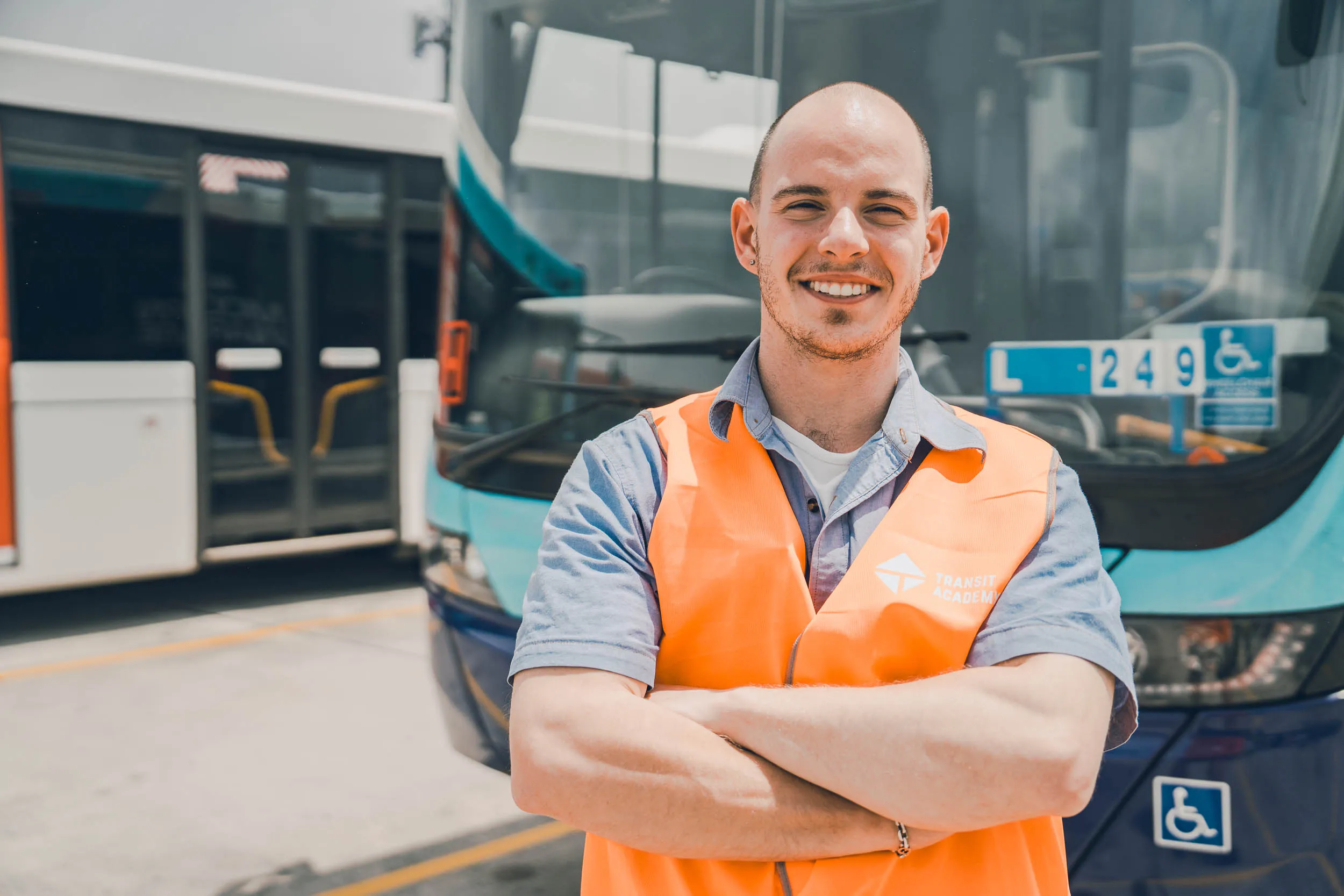 Smiling bus driver wearing an orange safety vest stands with arms crossed in front of buses.