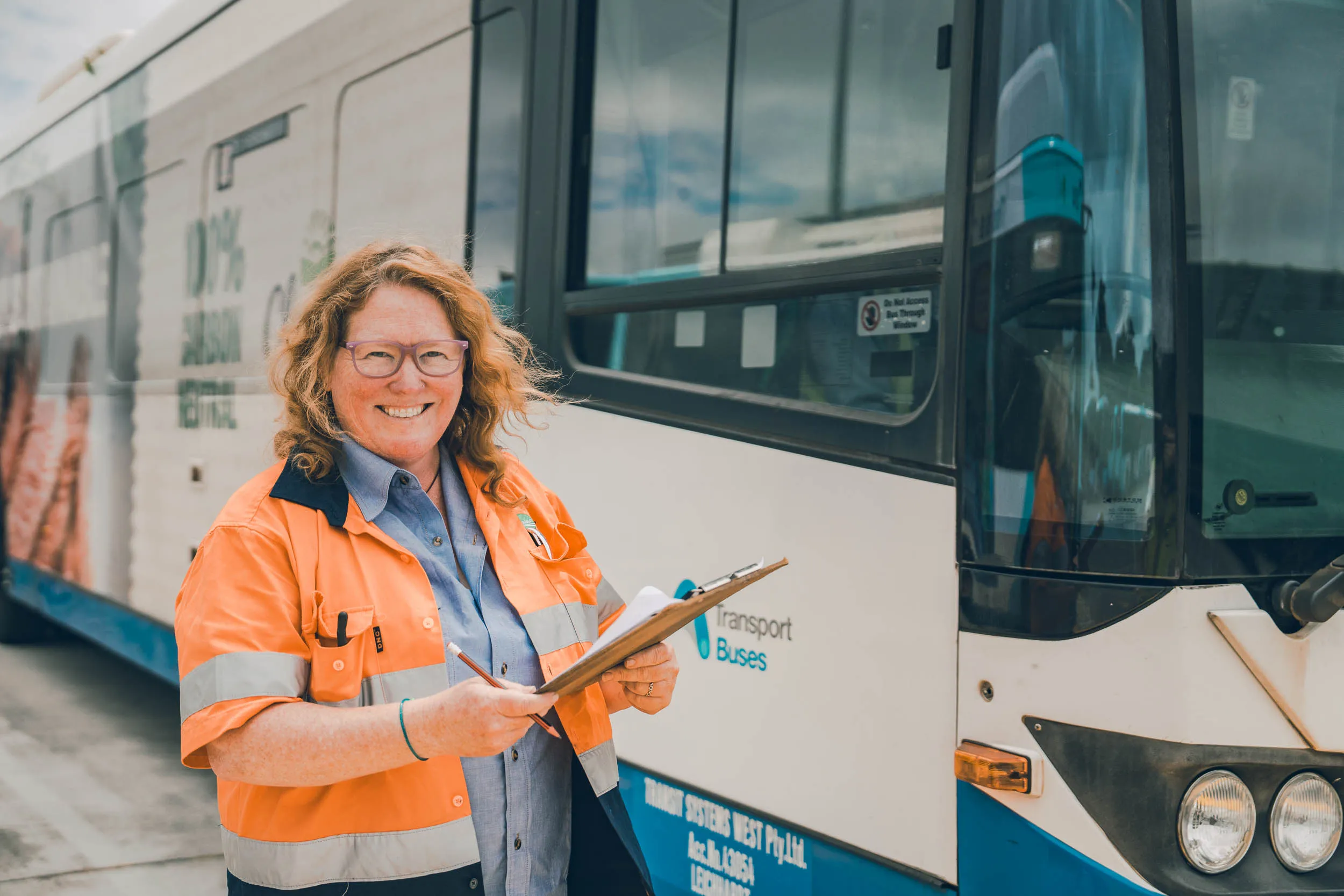 Woman in high-visibility jacket holding clipboard and pencil beside a public transport bus.