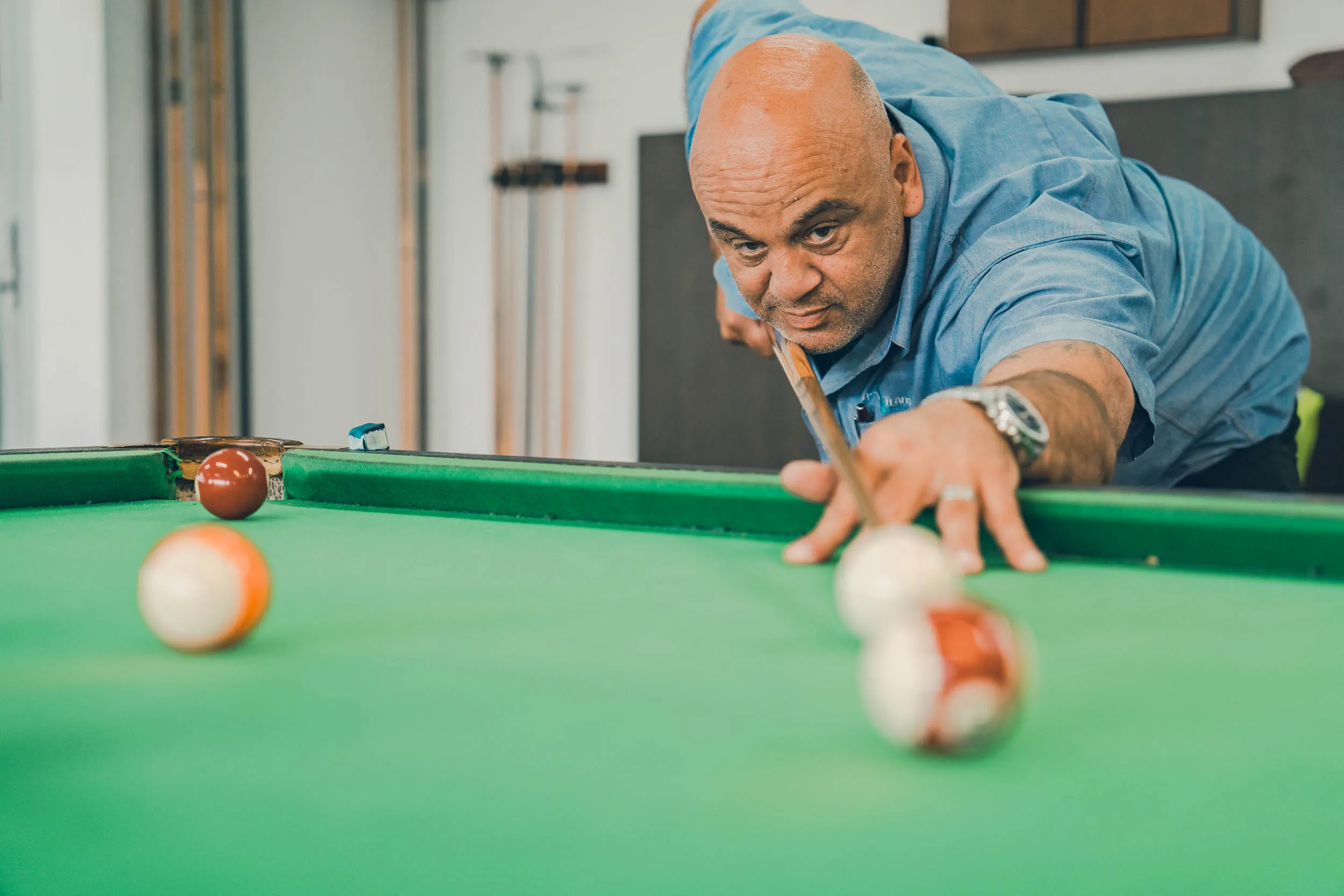 Man in blue shirt aiming with cue stick to play a shot on a green pool table.