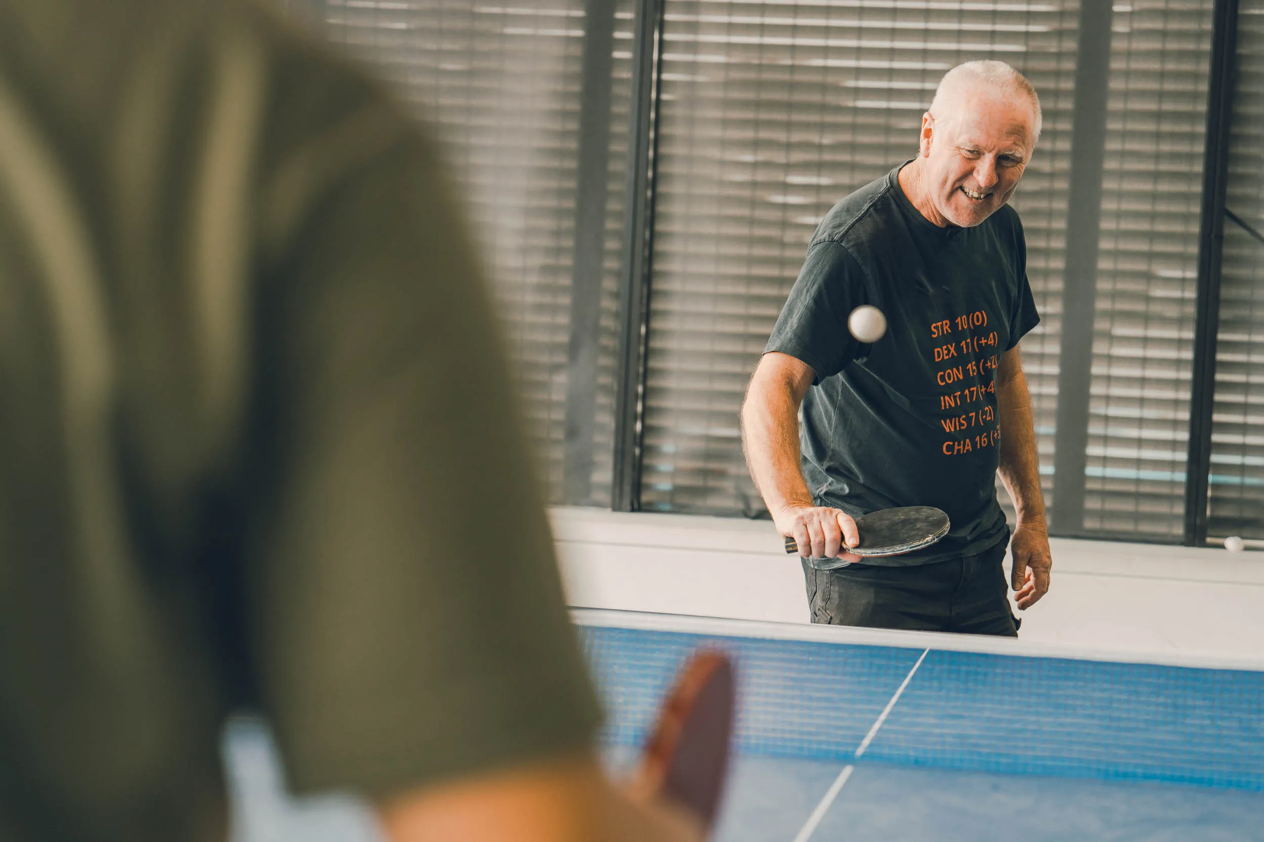 Smiling older man playing table tennis indoors, holding paddle and hitting the ball.