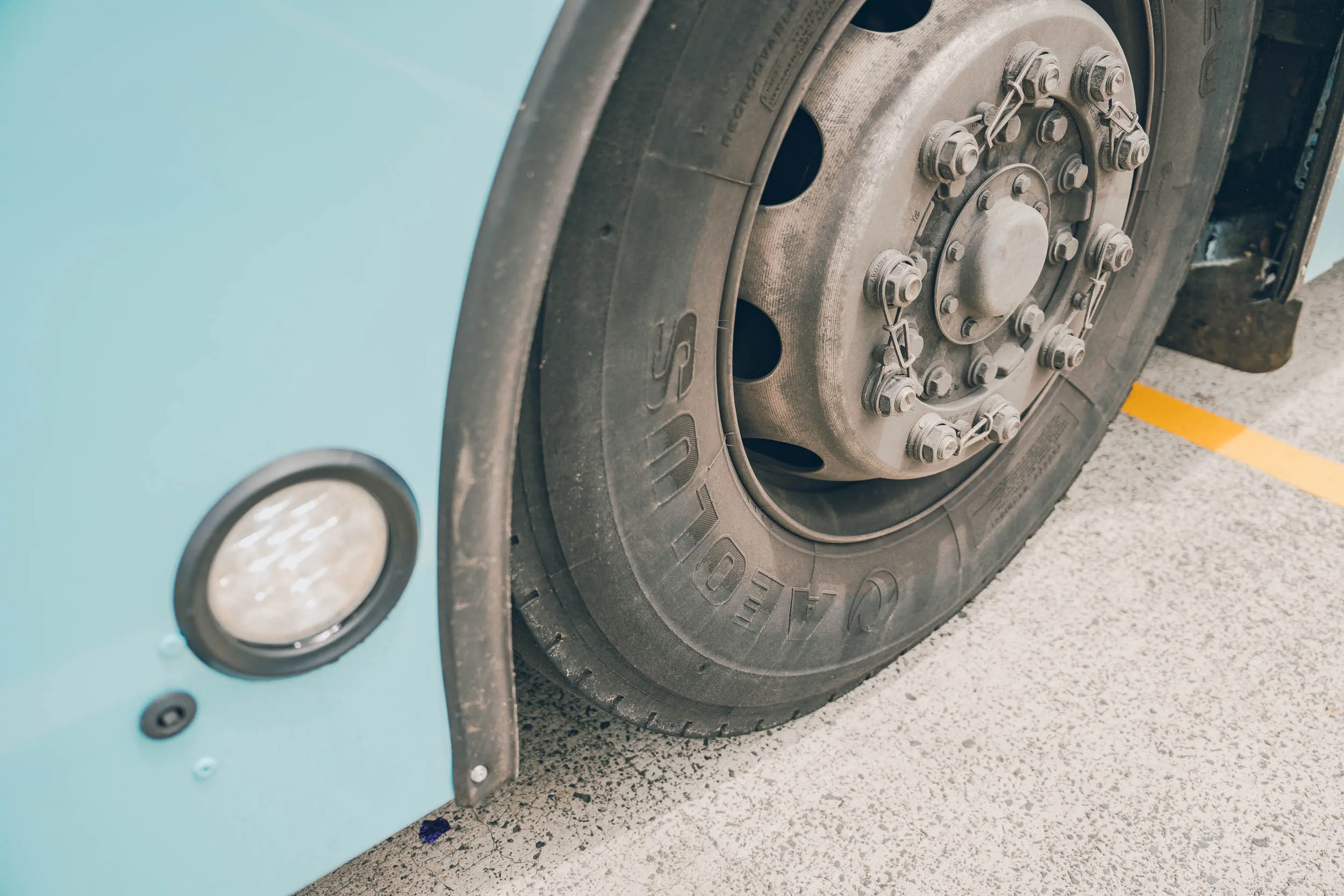 Close-up of large black bus tire on concrete near light blue vehicle body and yellow line.