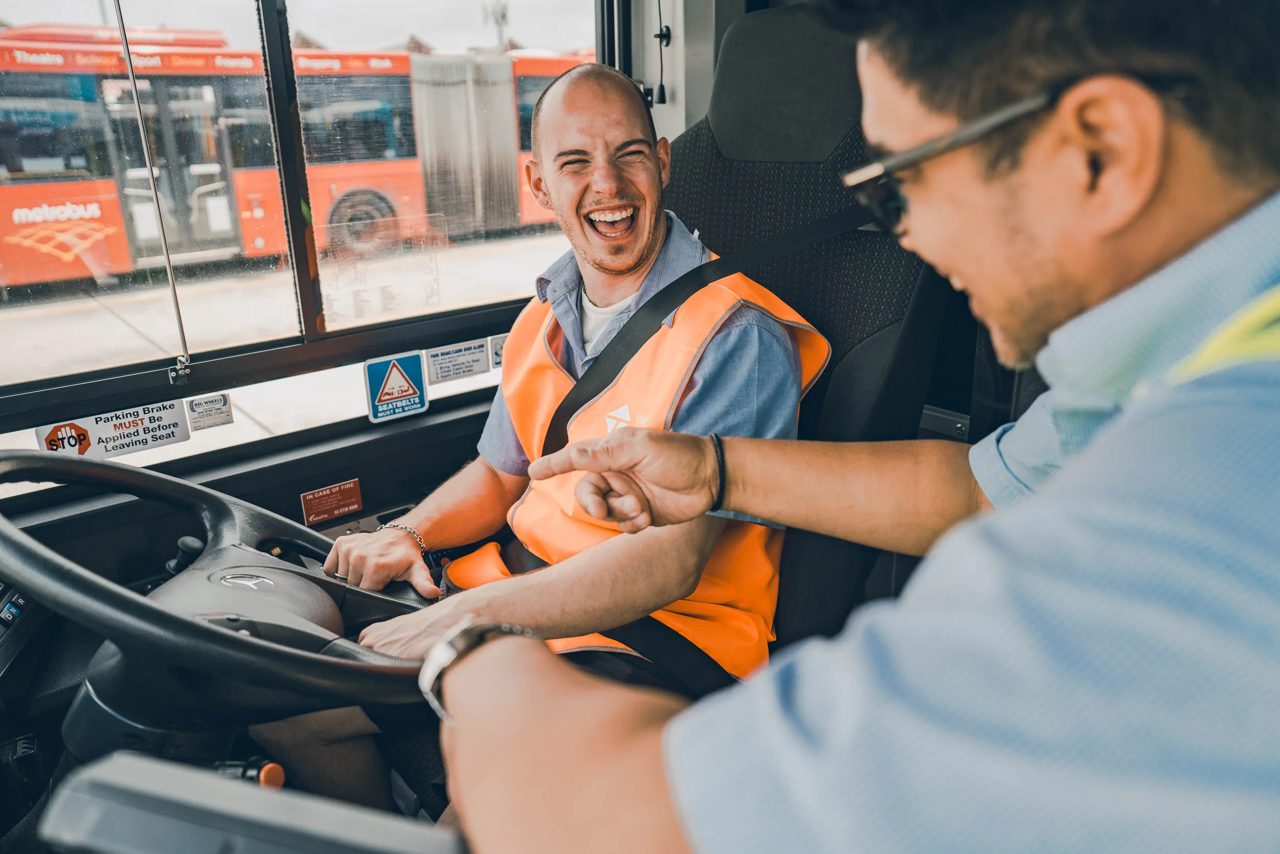Two men smiling inside a bus cabin, one in an orange vest holding the steering wheel.