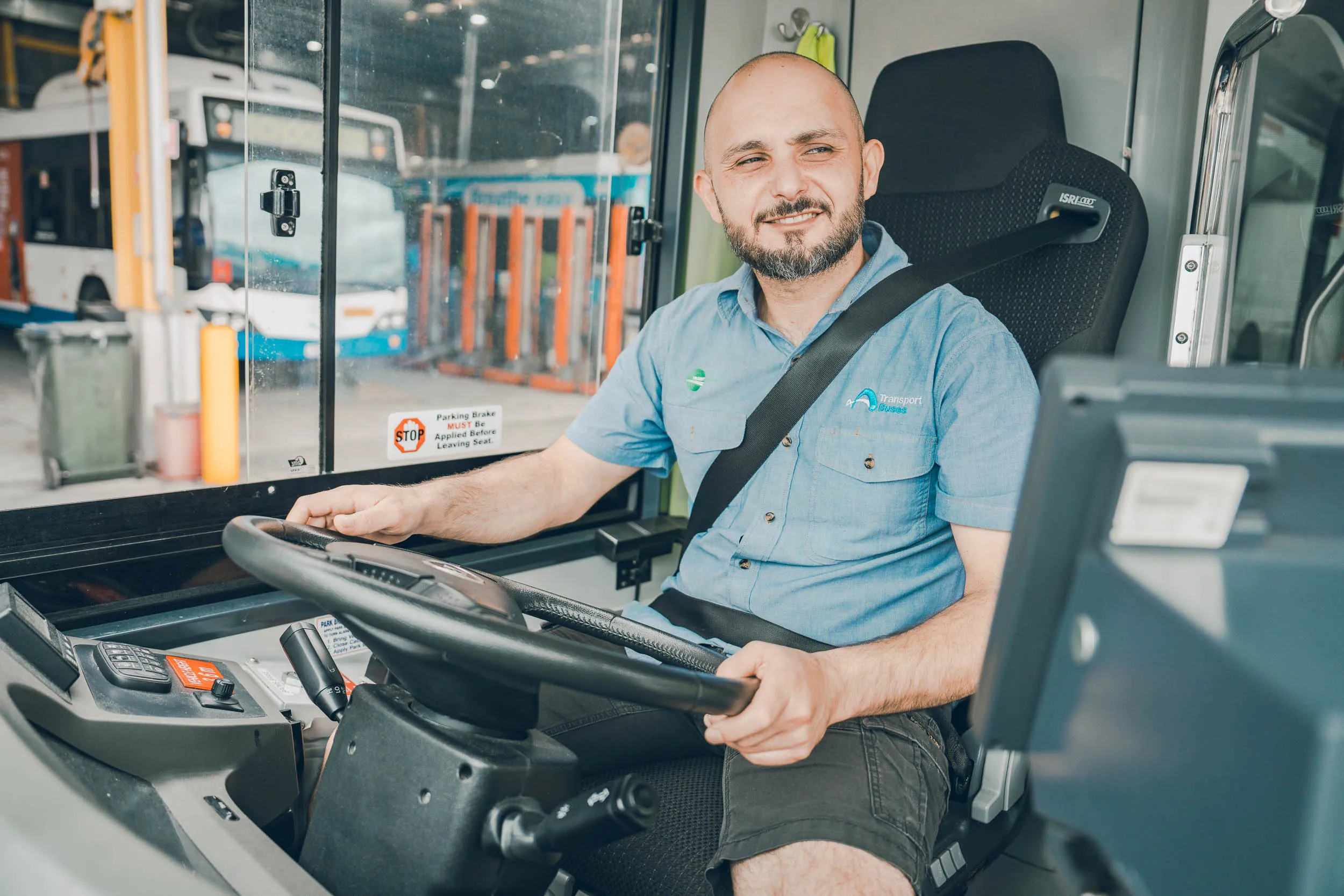 Smiling bus driver wearing seatbelt inside a bus cockpit with steering wheel and monitor visible.