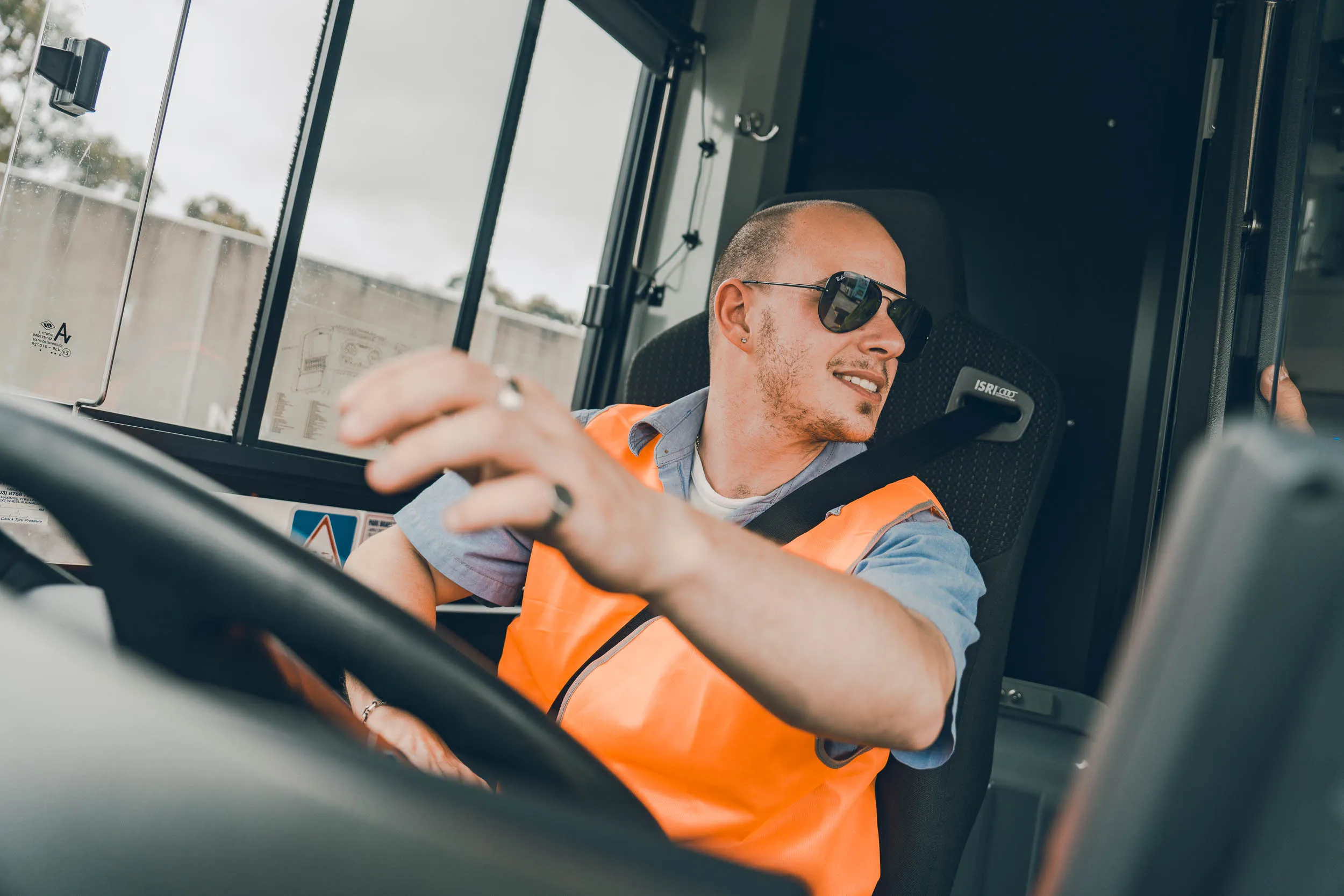 Man in orange safety vest and sunglasses driving a vehicle, looking to the side and smiling.