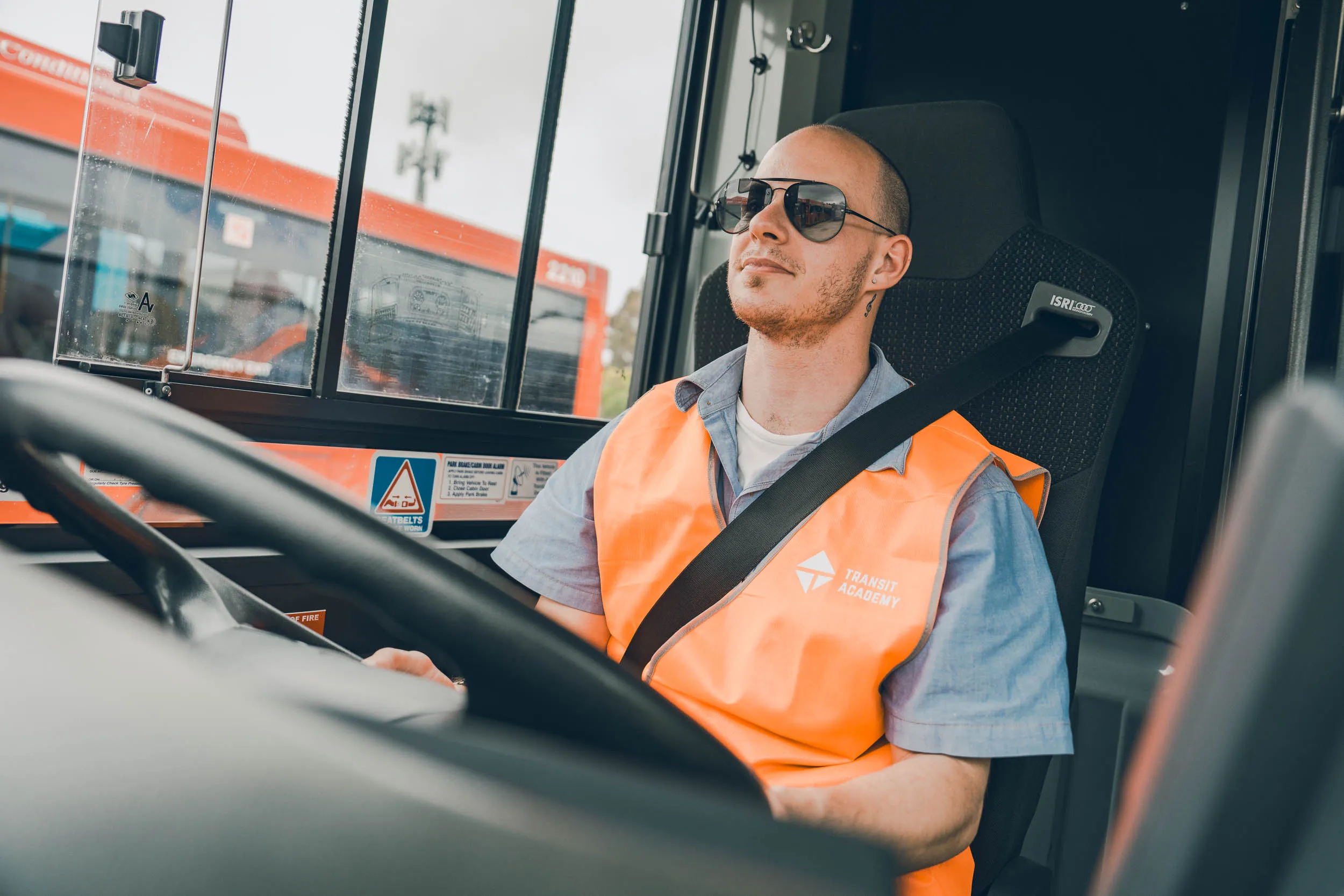 Bus driver wearing an orange Transit Academy vest and sunglasses, seated with seatbelt on.