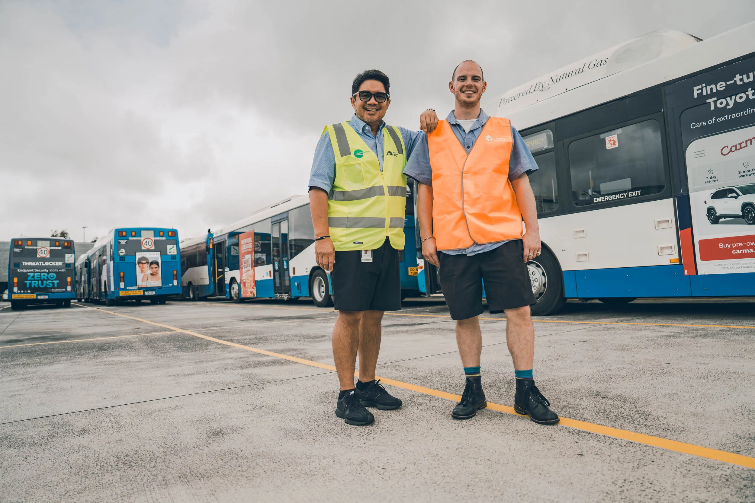 Two men wearing safety vests standing in front of parked blue and white buses on a cloudy day.