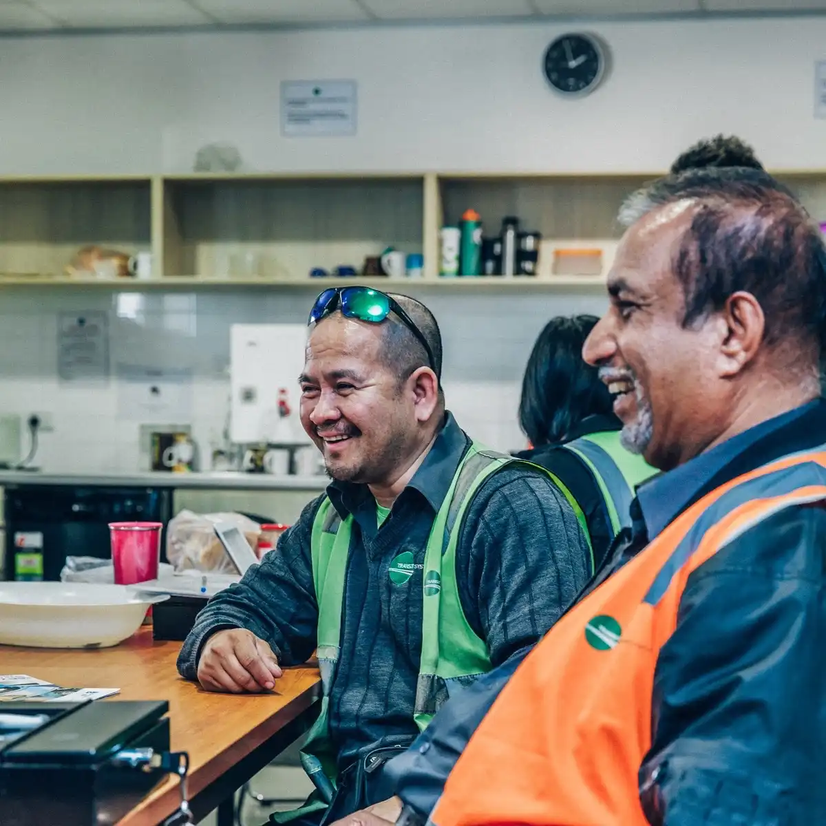 Two men in safety vests sitting at a table, smiling and chatting in a break room.
