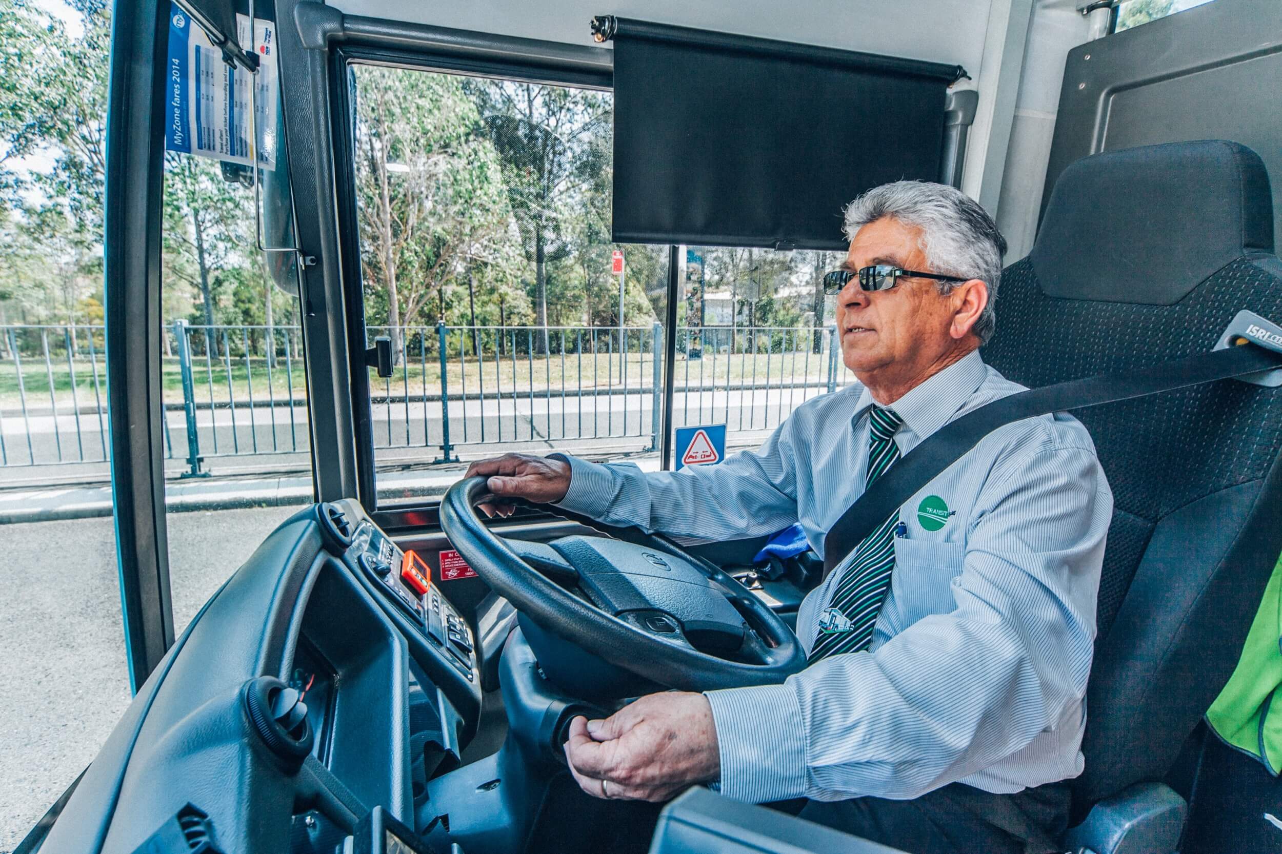 Older male bus driver wearing sunglasses and uniform driving a bus with seatbelt on.