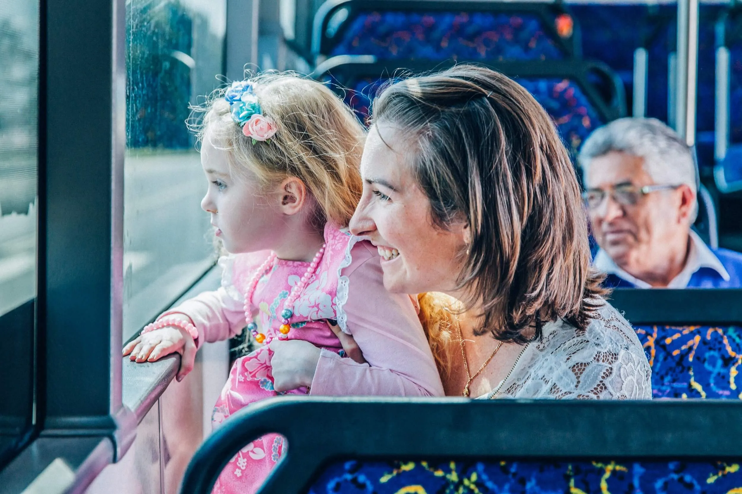Smiling woman holding a young girl looking out the window on a colorful bus seat.