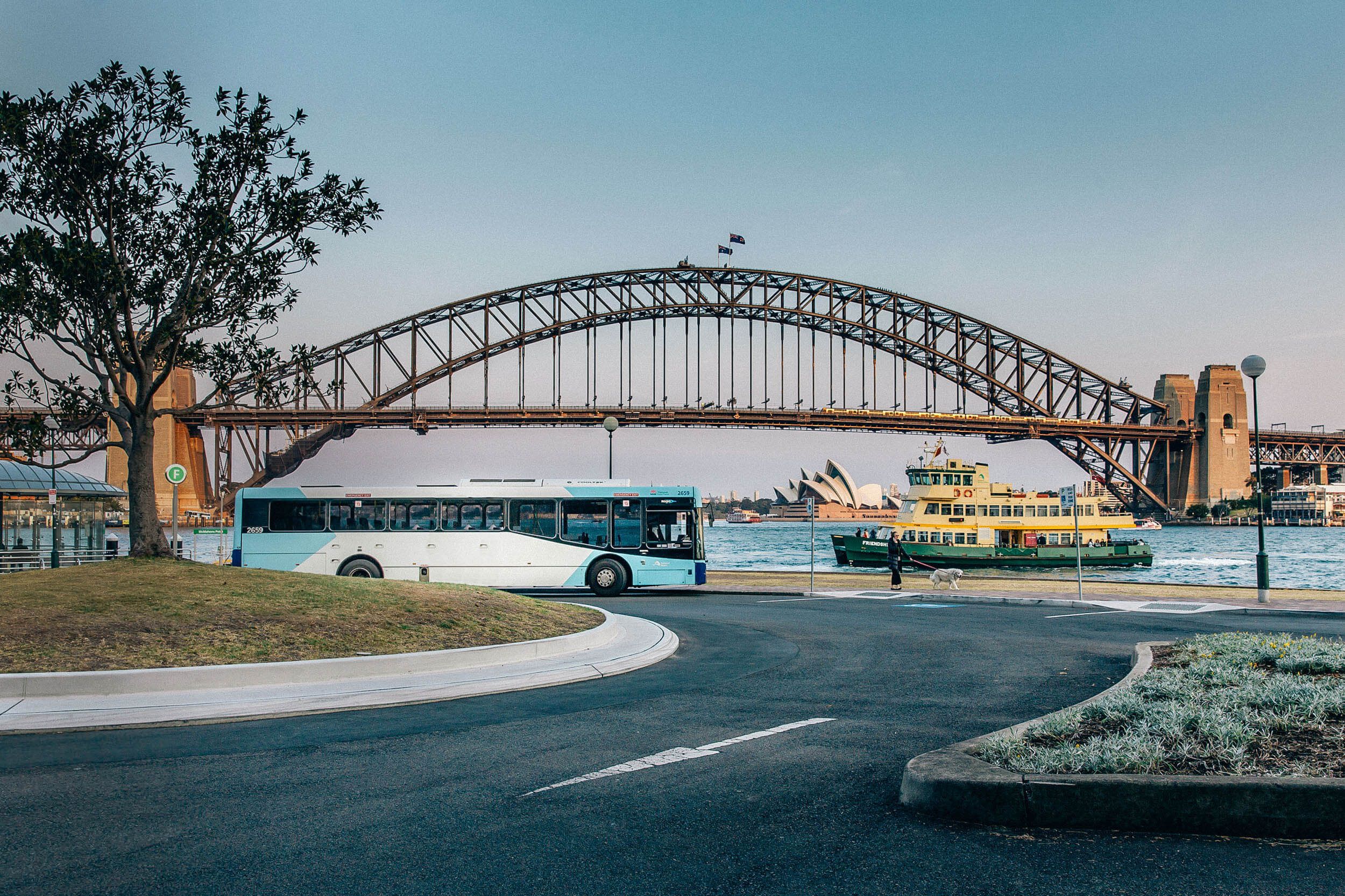 Sydney Harbour Bridge with a bus, ferry, and Sydney Opera House in the background at sunset.