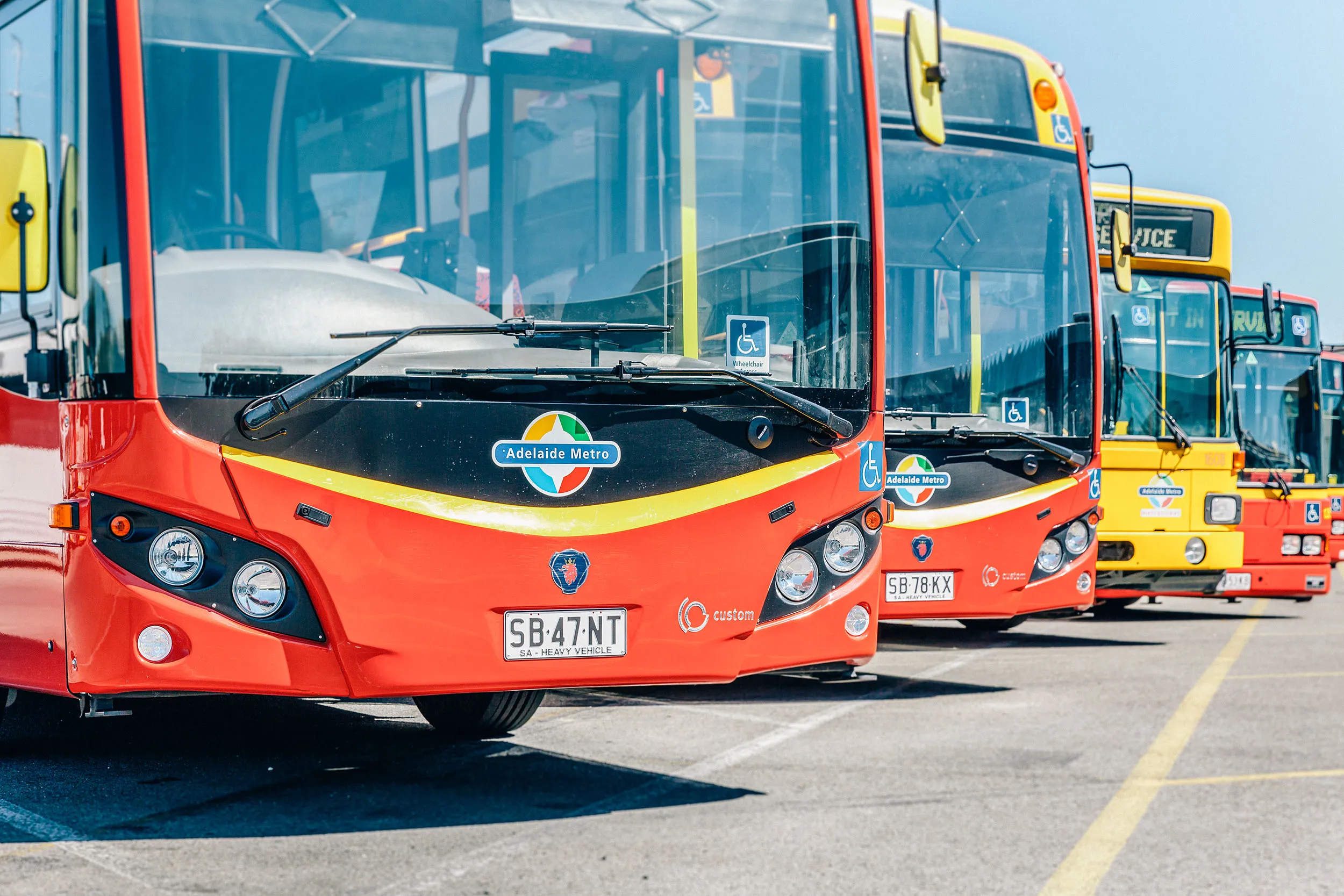 Row of red and yellow Adelaide Metro buses parked in a bus depot under clear sky.