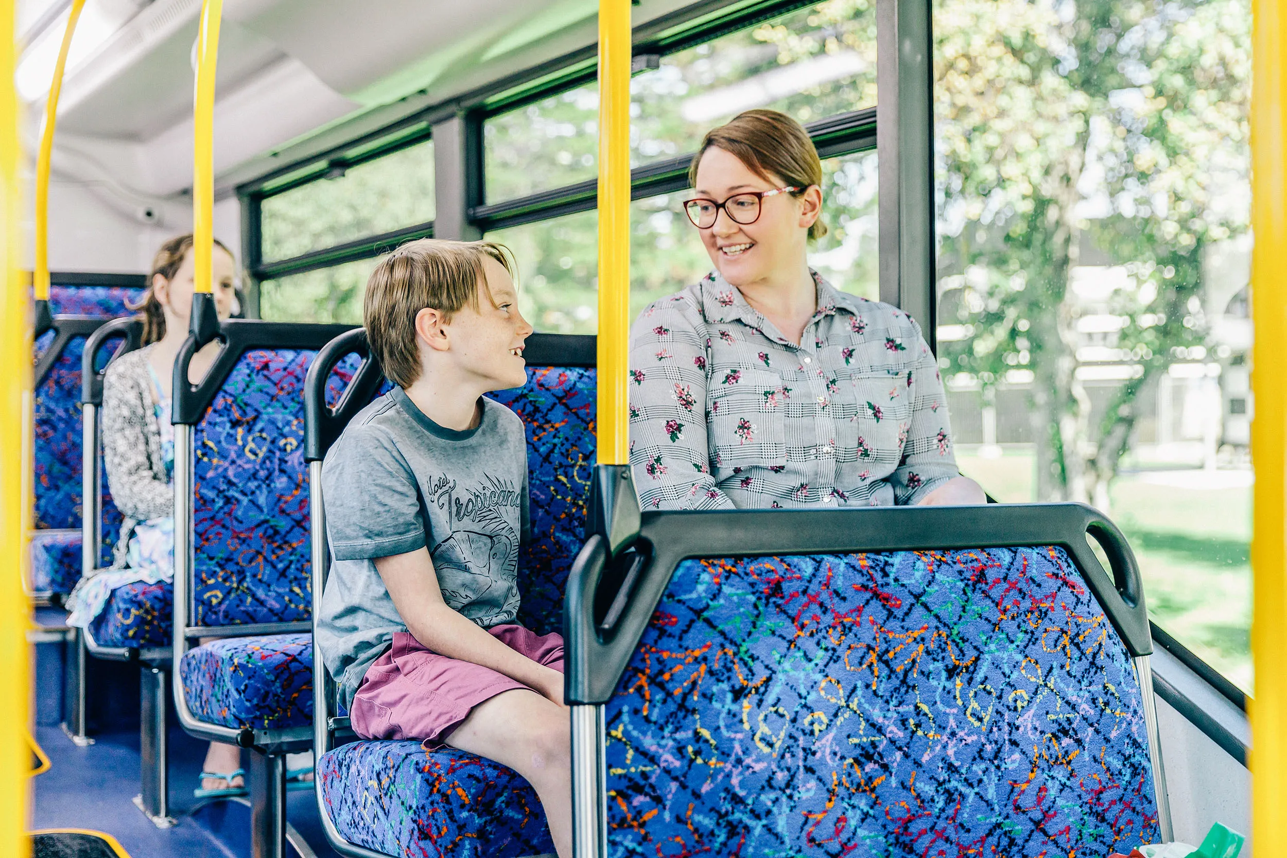Woman wearing glasses smiling and talking with a boy in pink shorts on a bus with colorful seats.