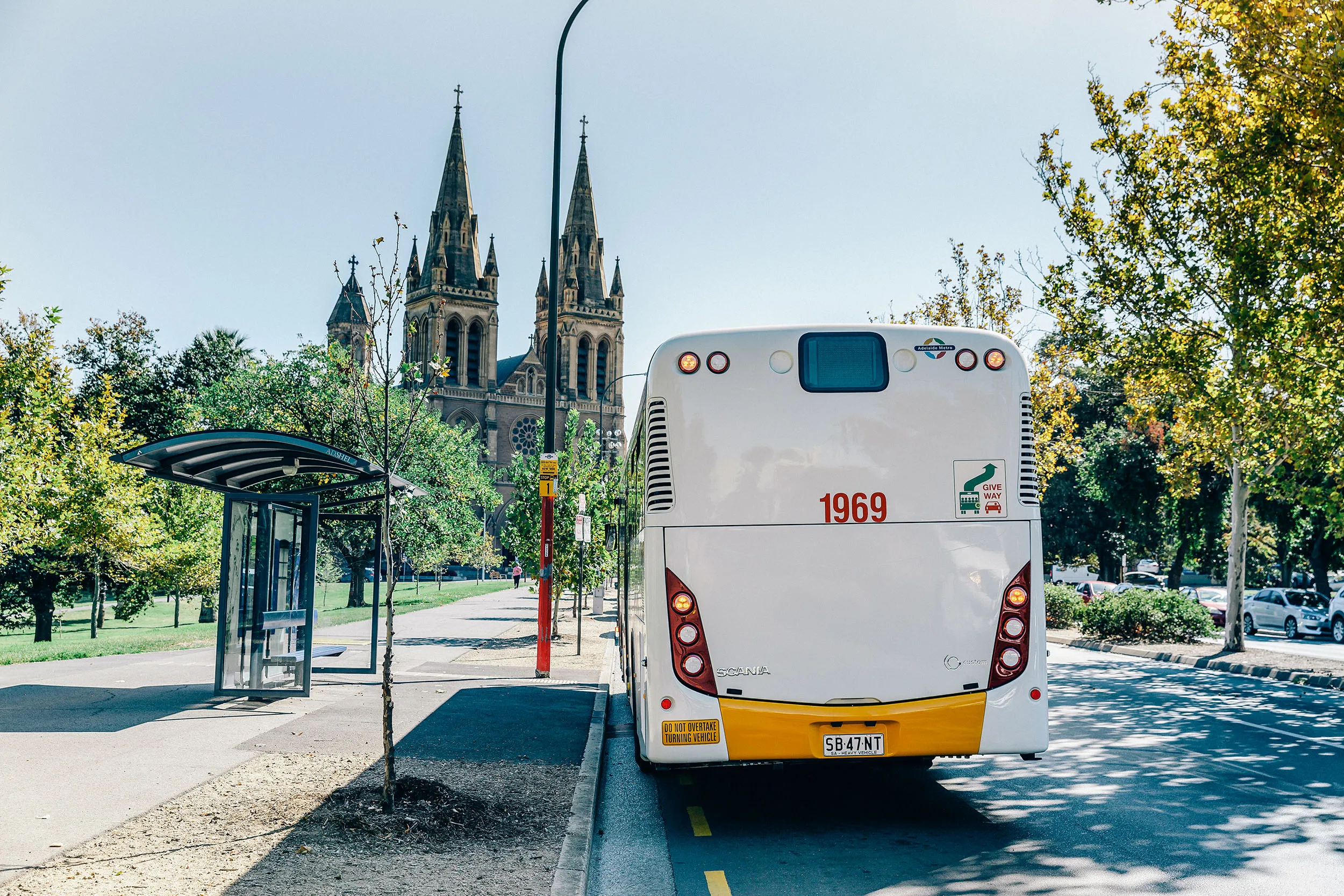 Rear view of white Adelaide Metro bus parked by a shelter near trees and a Gothic-style cathedral.