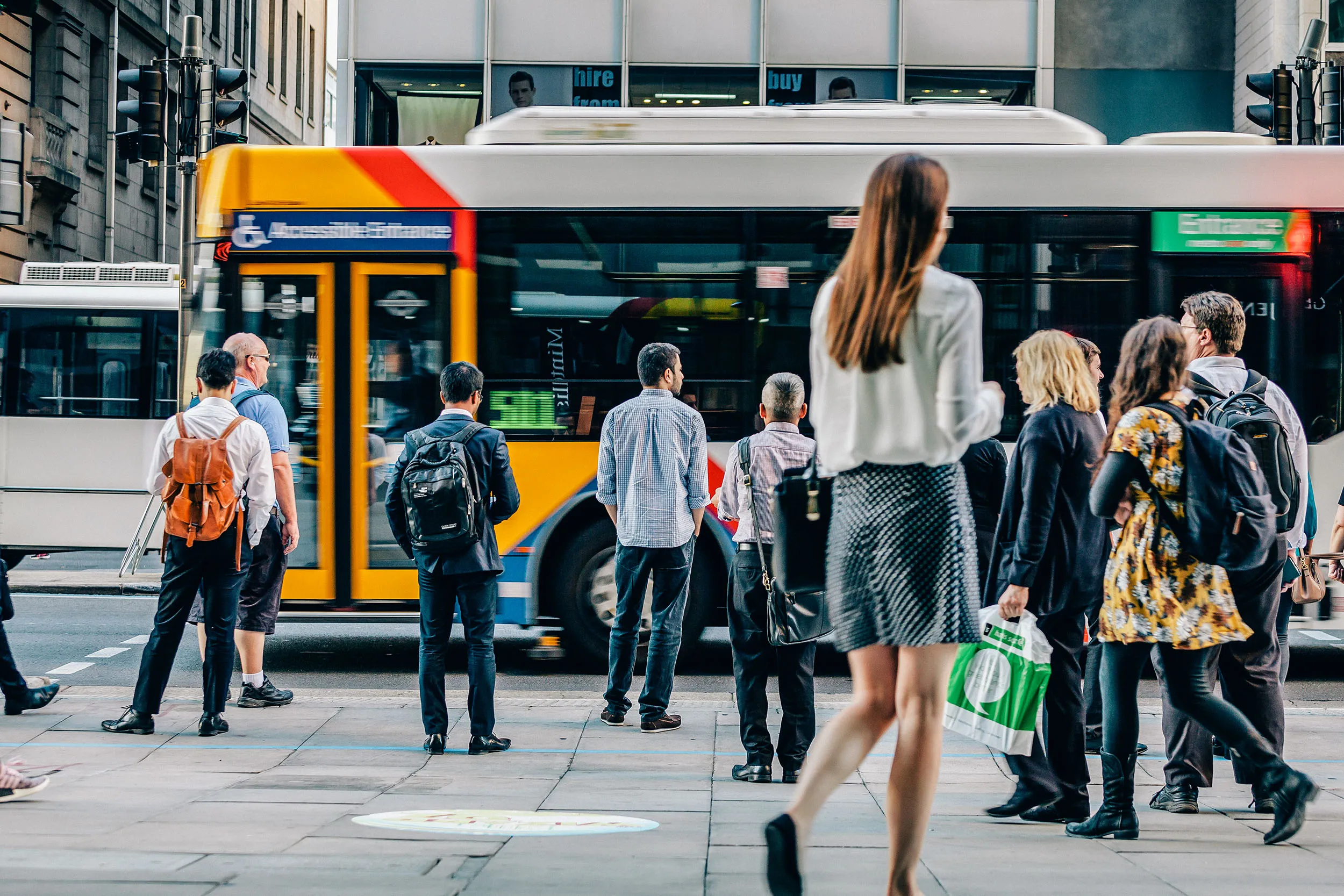 People waiting at a busy city bus stop with a yellow and white bus passing by in the background.