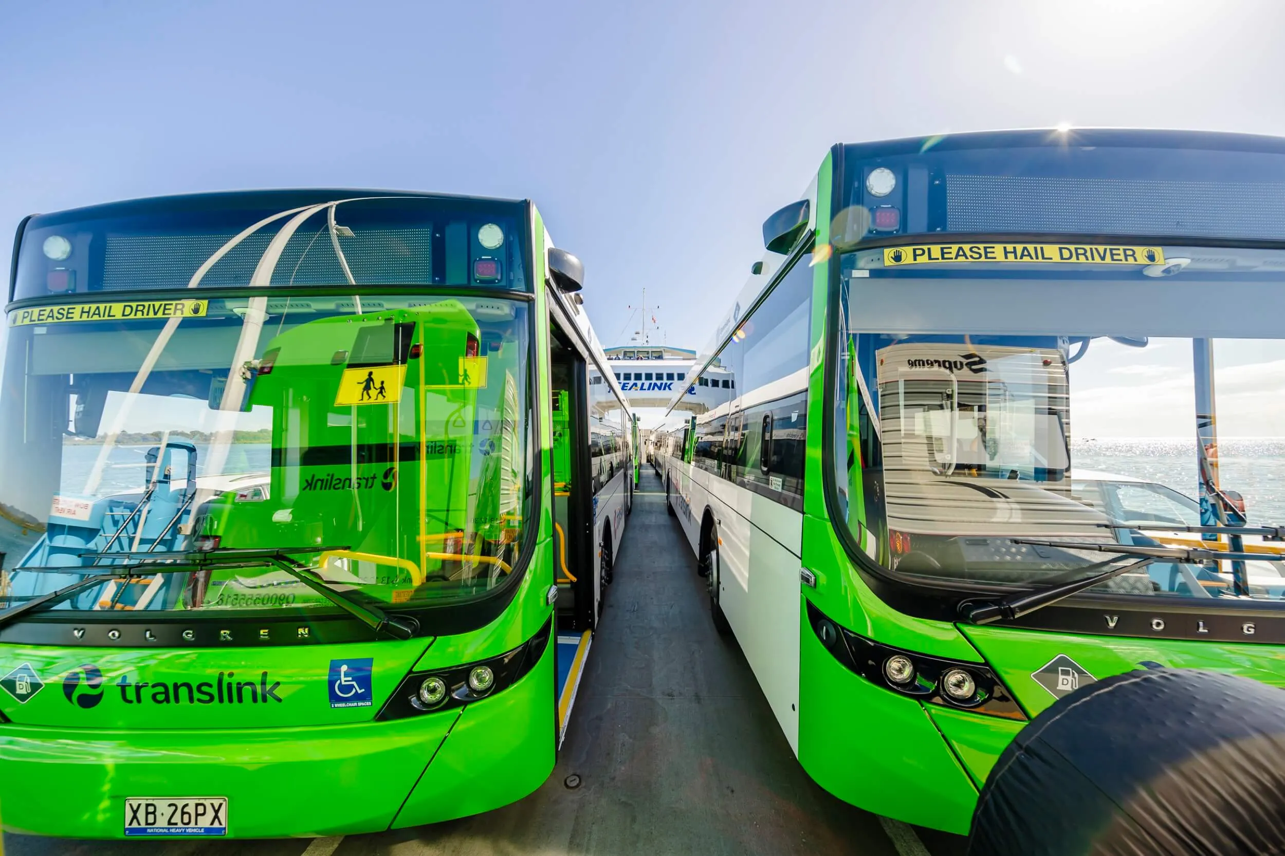 Two green Translink buses parked side by side on a ferry under a clear blue sky.