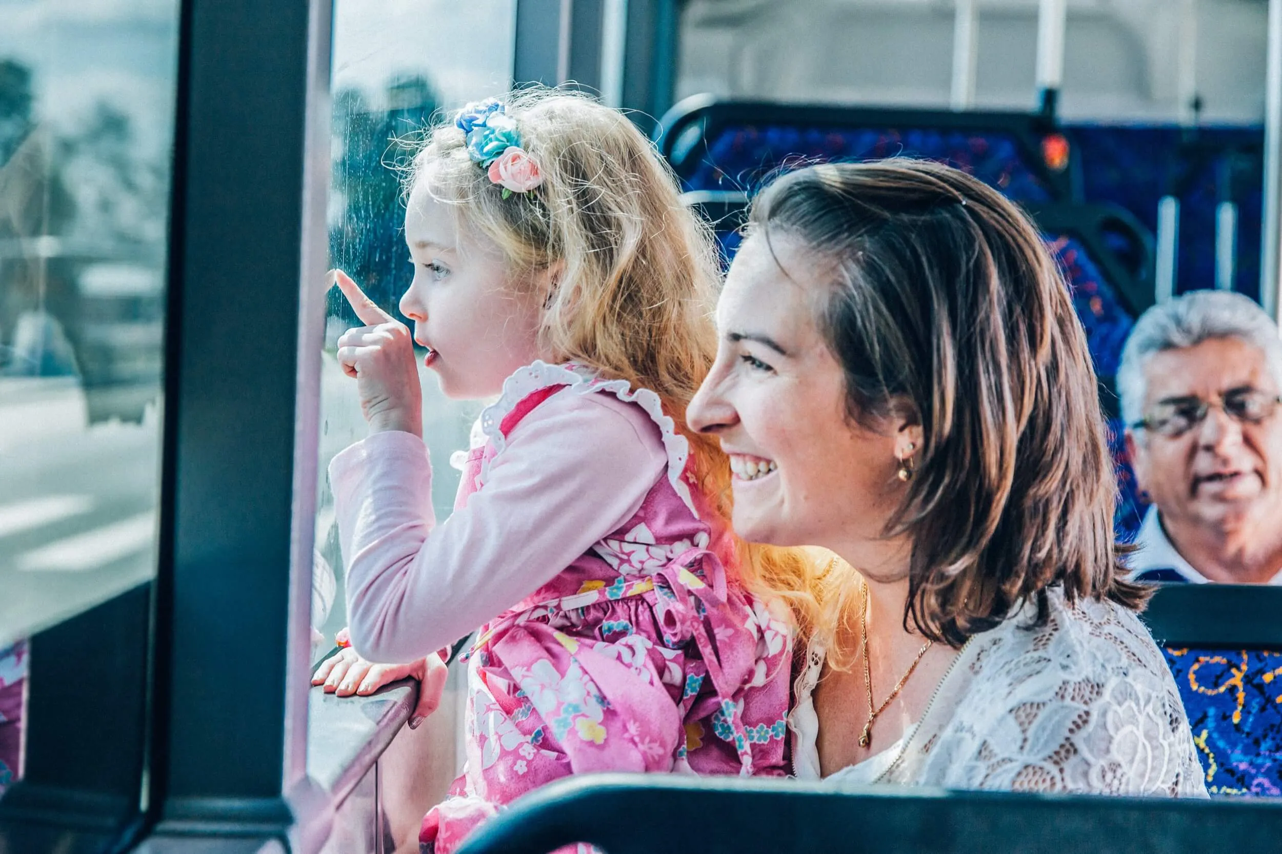 Smiling woman holding girl in floral dress pointing outside window on a bus.