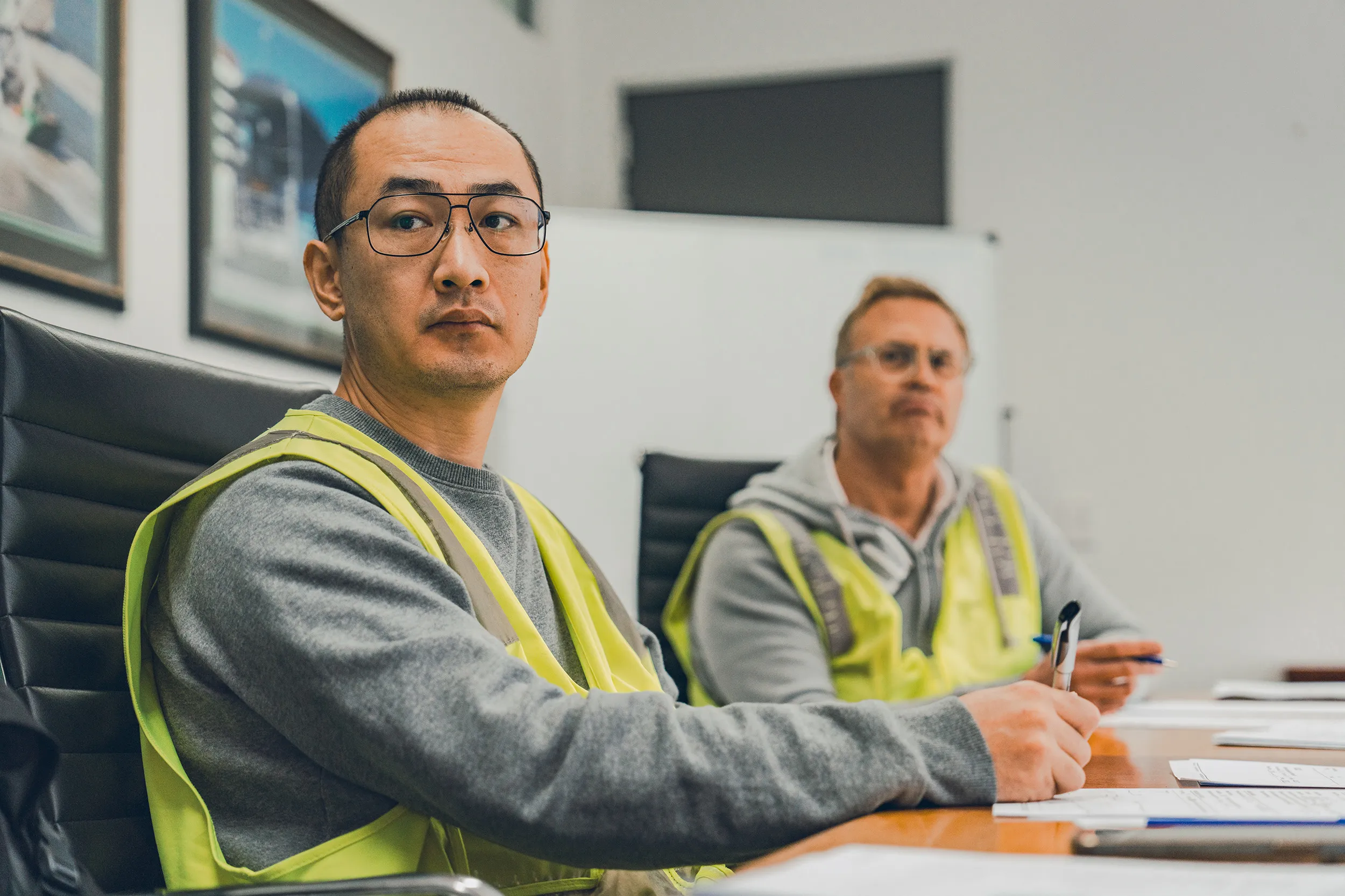 Two men wearing yellow safety vests sitting at a conference table with papers and pens.