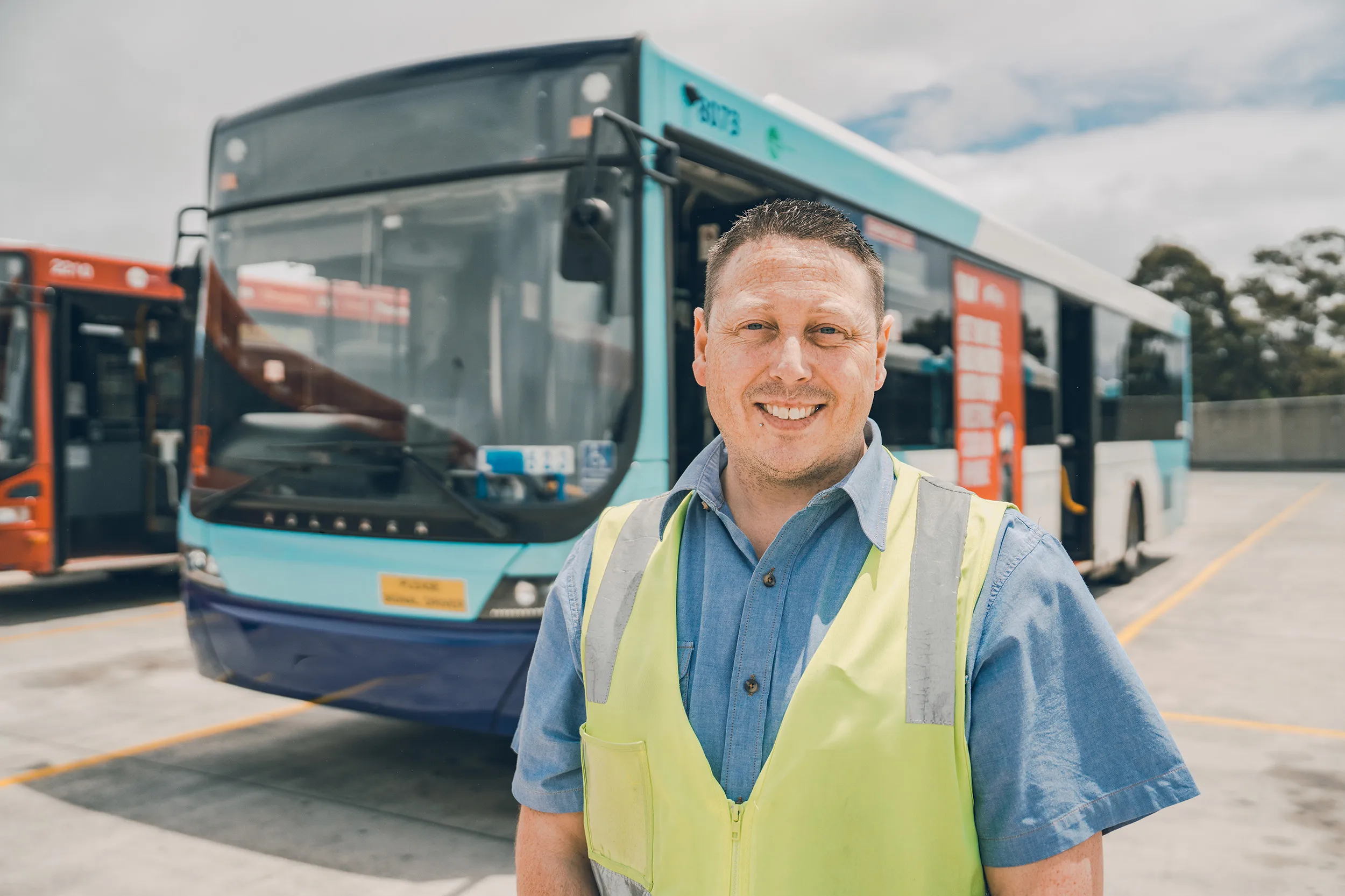 Smiling man in a safety vest stands in front of parked transit buses at a depot.