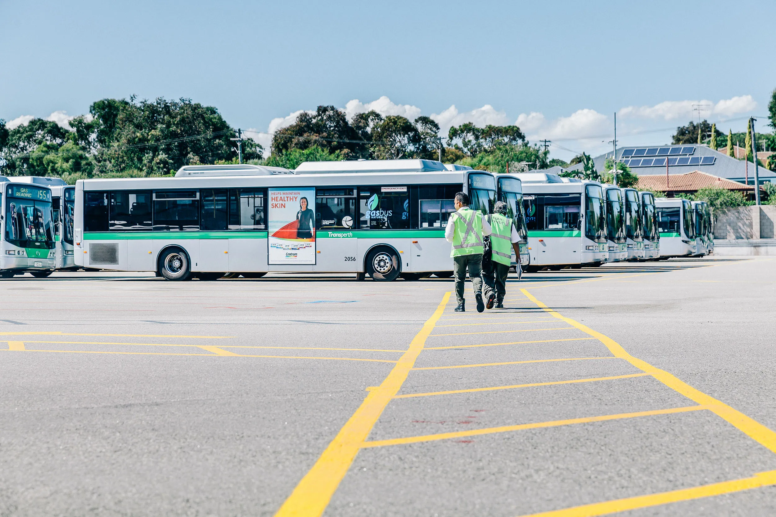 Two workers in reflective vests walk toward a row of parked Transperth buses on a sunny day.