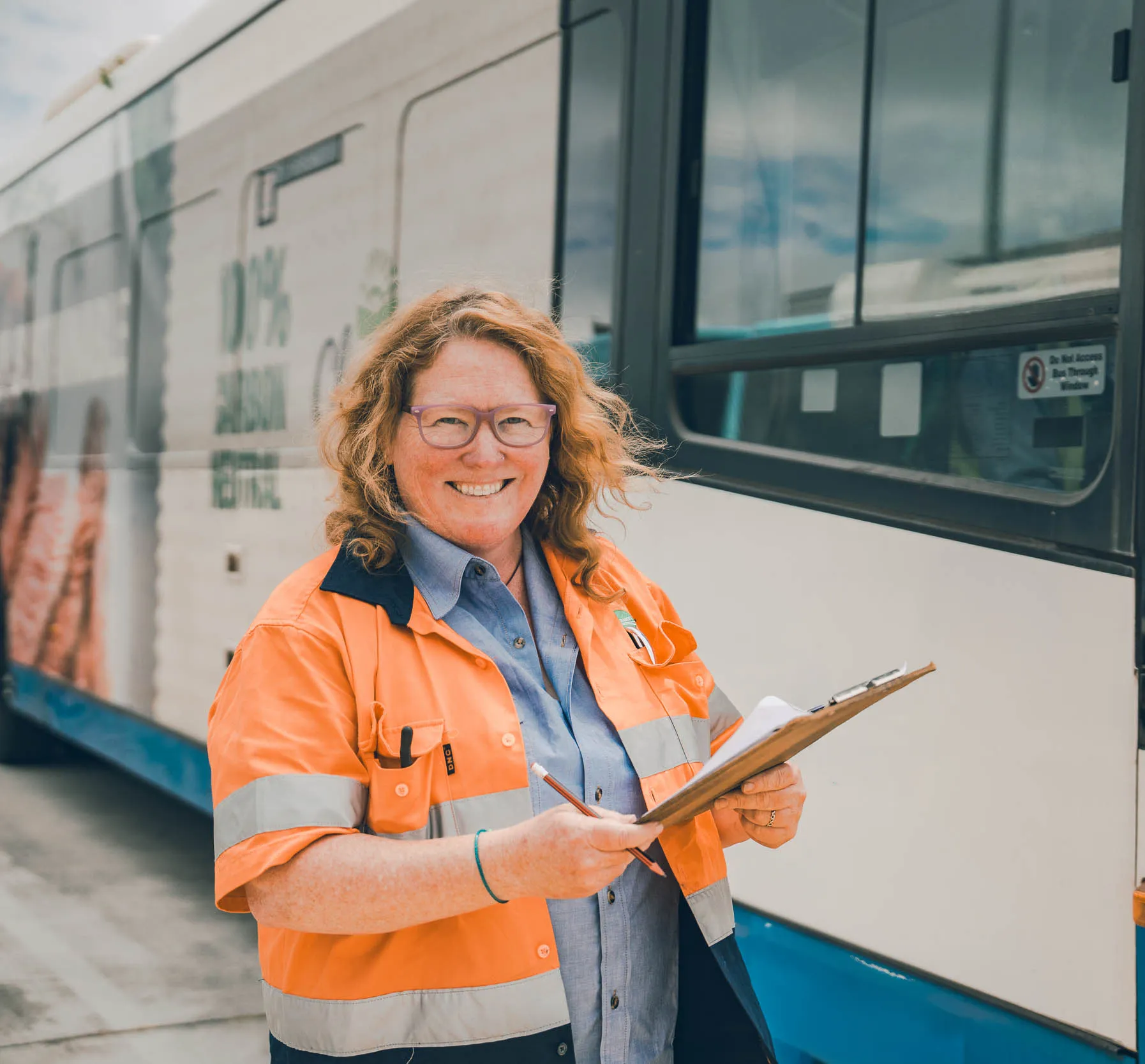 Smiling woman in orange safety jacket and glasses holds clipboard next to a bus.
