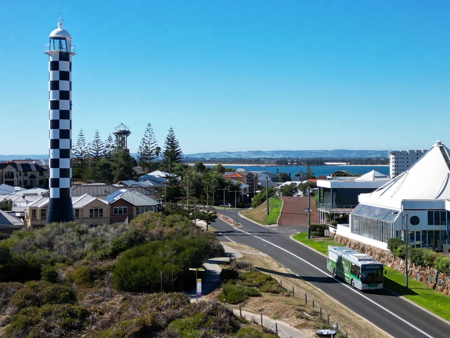 Black and white checkered lighthouse near coastal houses with a bus on the road under a clear blue sky.