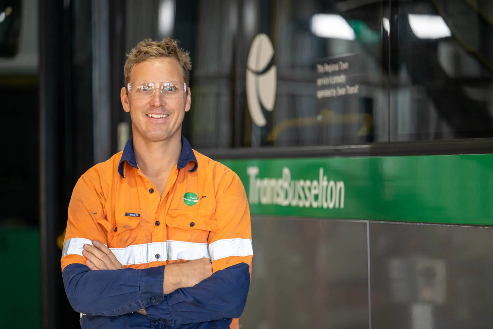 Smiling man in orange and blue workwear with safety glasses stands in front of a TransBusselton bus.