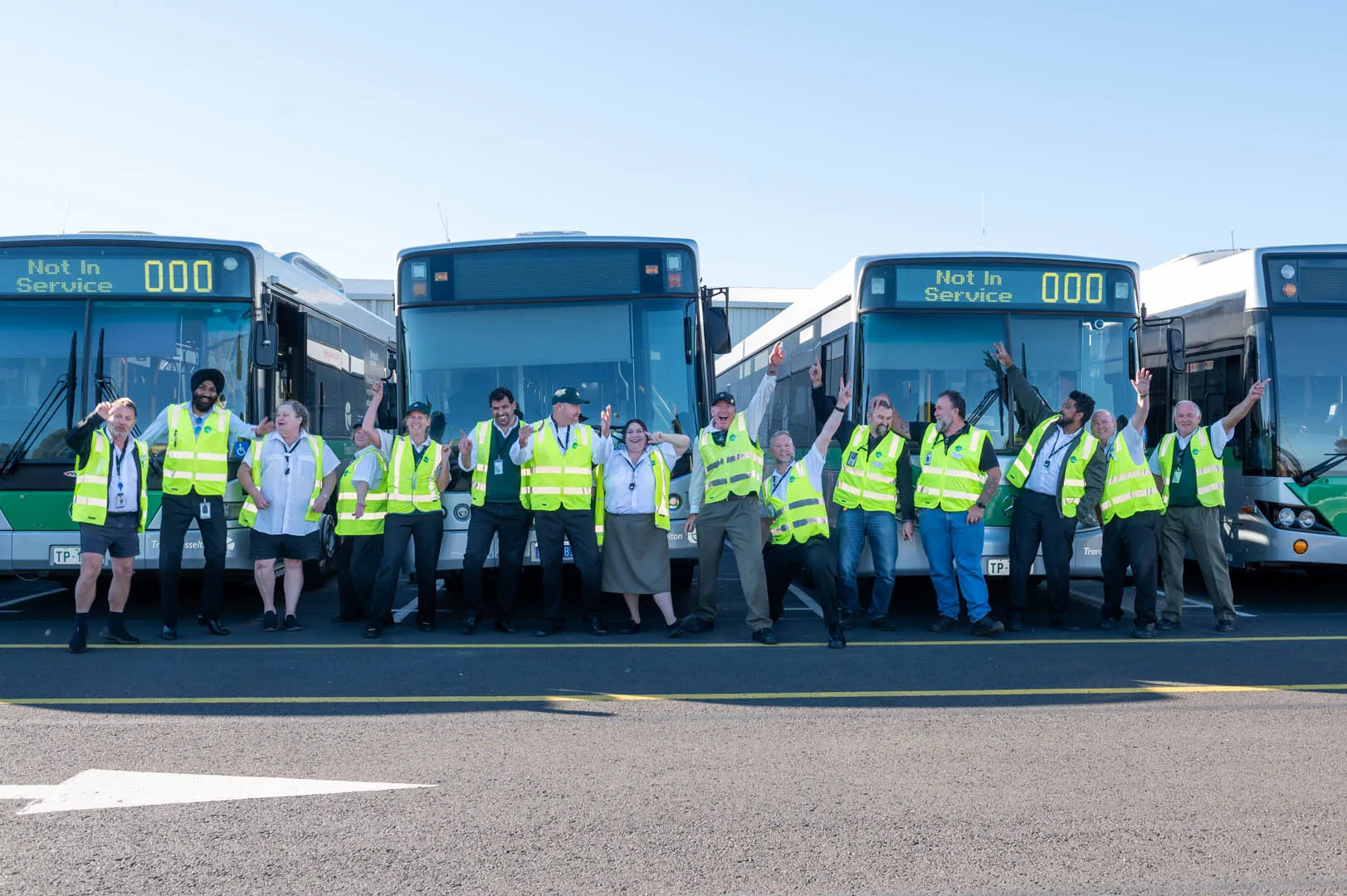 Group of transit workers wearing reflective vests standing and posing happily in front of parked buses.