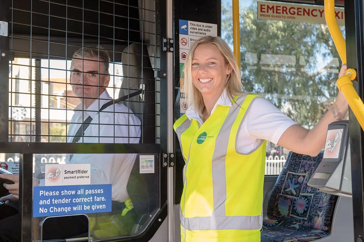 Smiling female transit worker in reflective vest standing at bus door next to seated male driver.