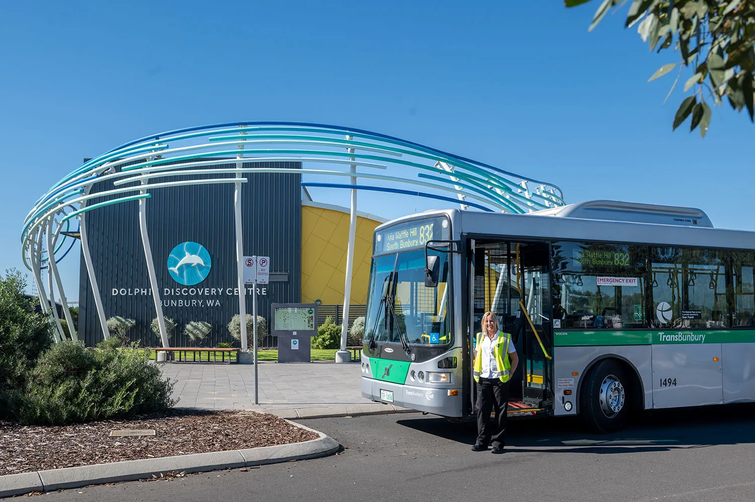 TransBunbury bus with driver outside Dolphin Discovery Centre in Bunbury, WA under a clear blue sky.