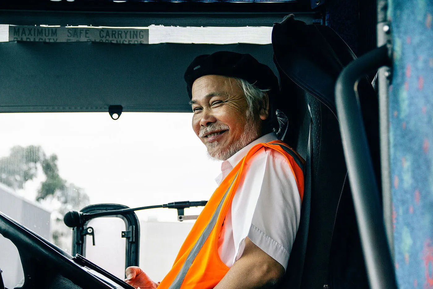 Smiling older man wearing an orange safety vest and black beret sitting in a vehicle driver's seat.