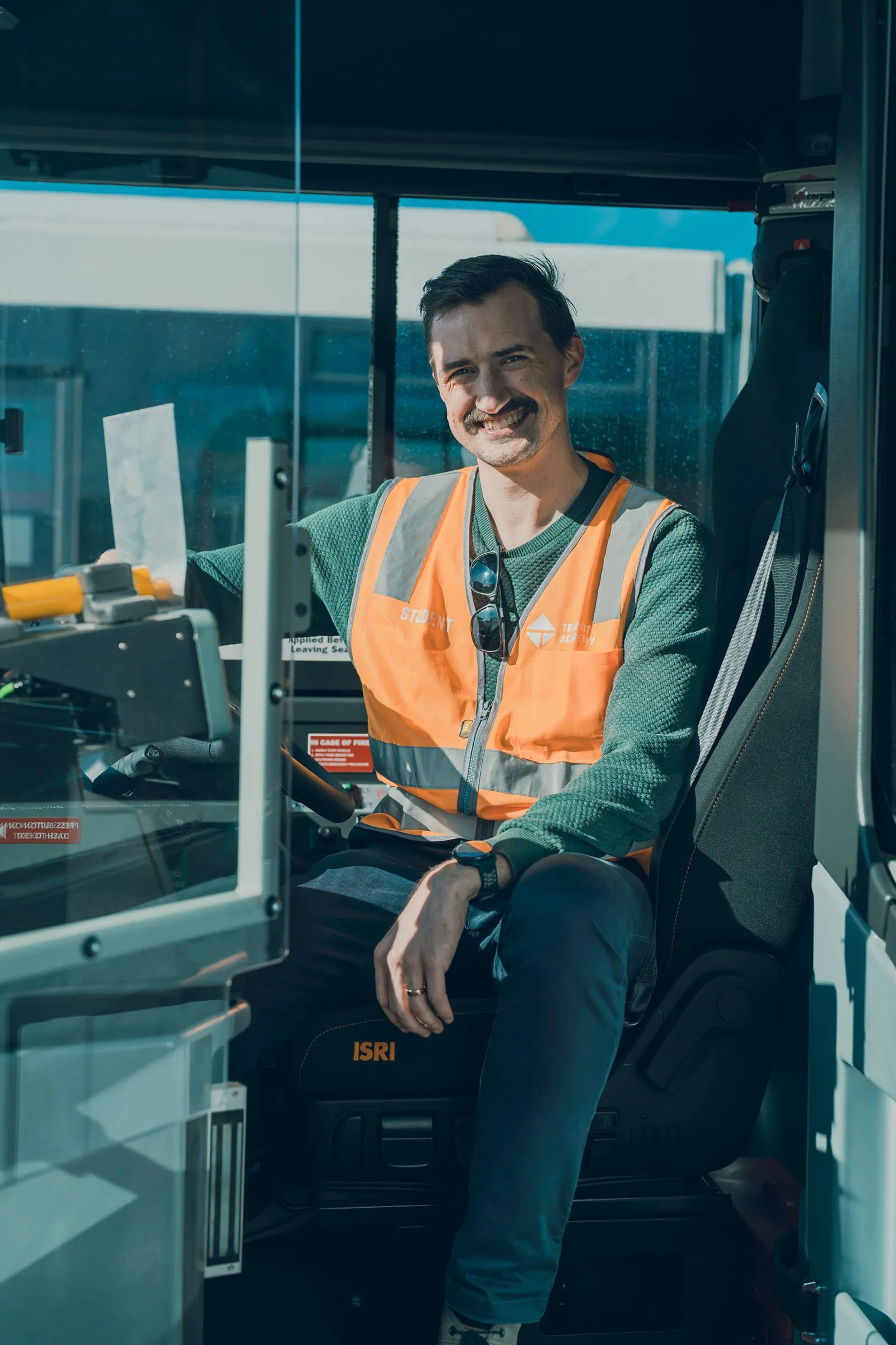 Smiling man wearing an orange safety vest sitting in a vehicle driver's seat with a steering wheel.