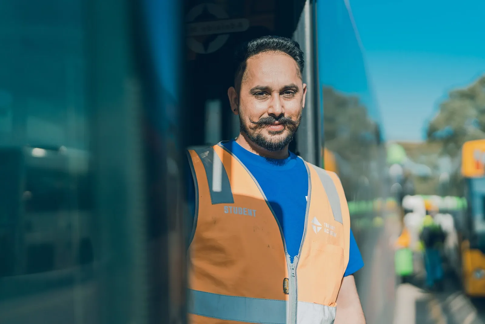 Man in blue shirt and orange safety vest labeled Student stands next to bus on sunny day.