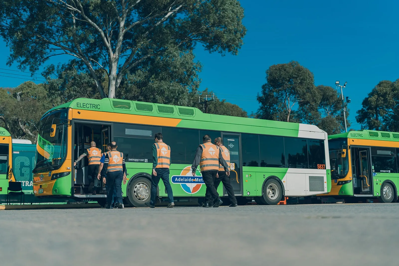 Five people in orange safety vests board a green and yellow electric Adelaide Metro bus outdoors.