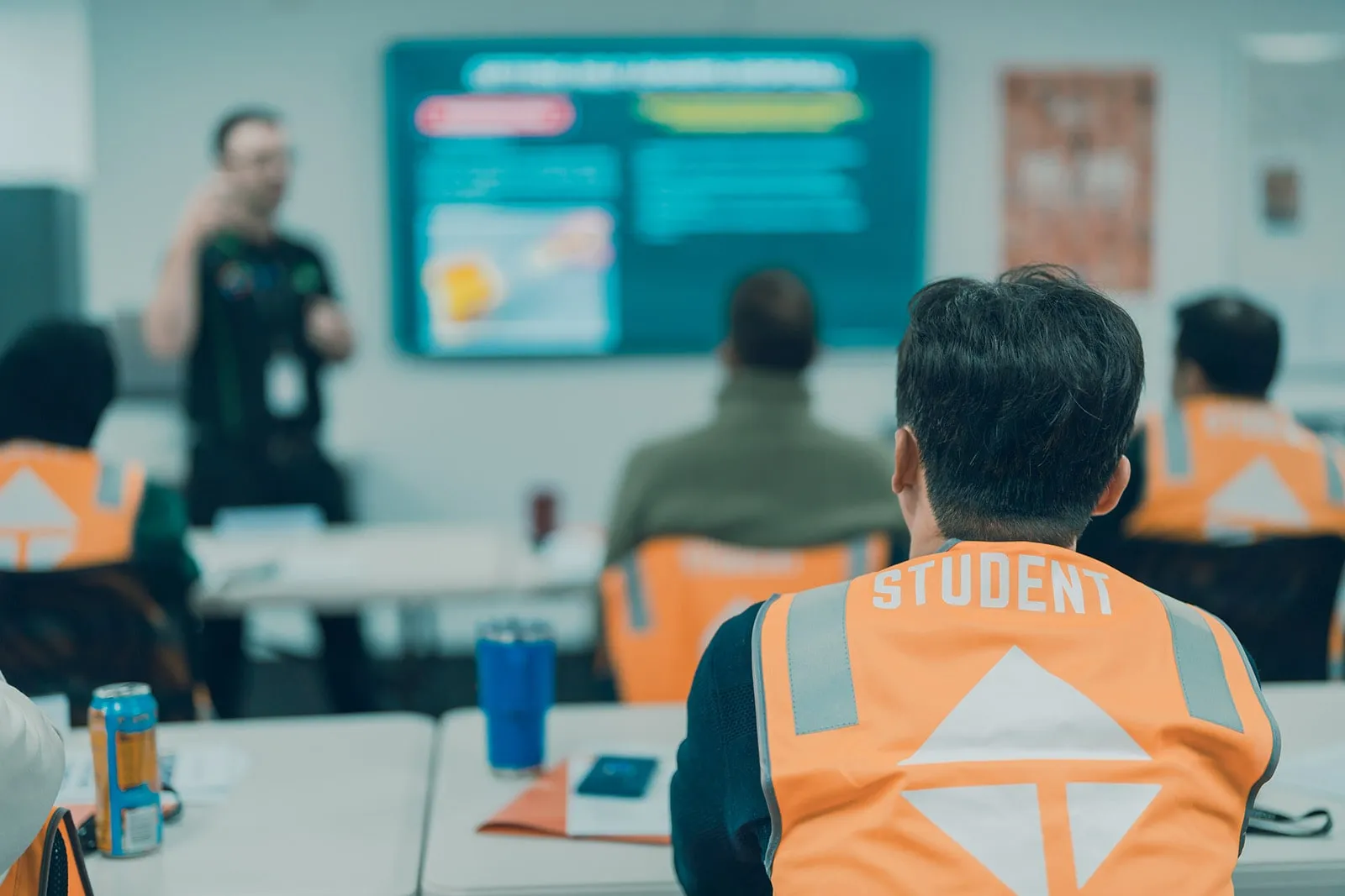 Students wearing orange safety vests attend a classroom training with a presenter at the front.