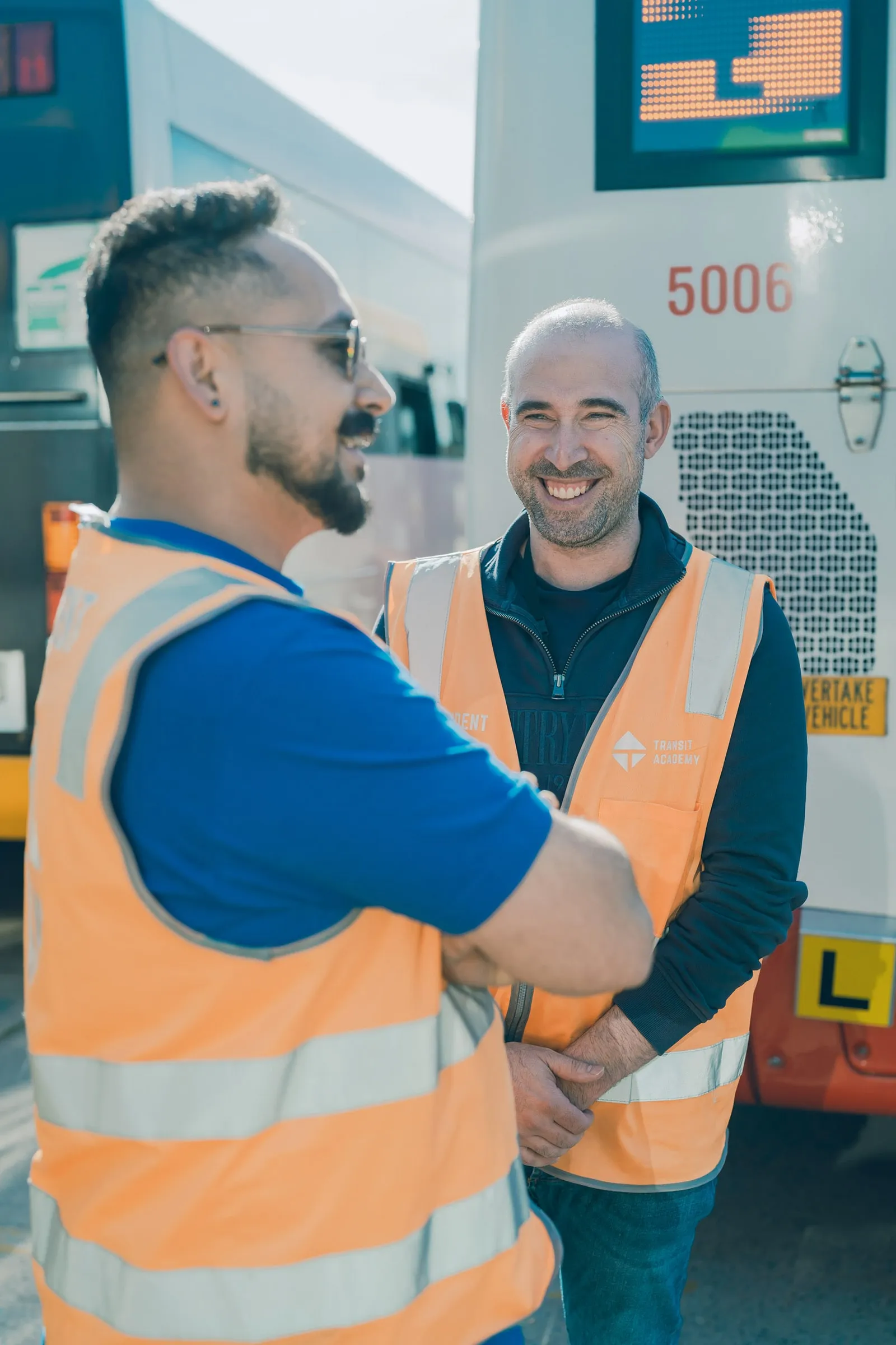 Two men in orange safety vests smiling and talking near buses parked outside.