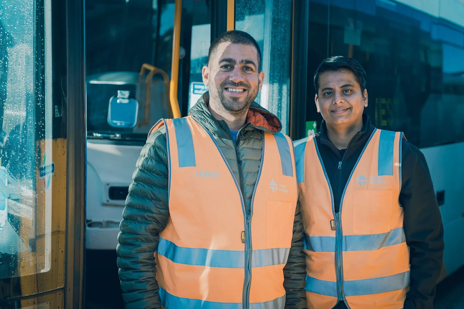 Two men wearing orange safety vests standing beside a bus with smiles.