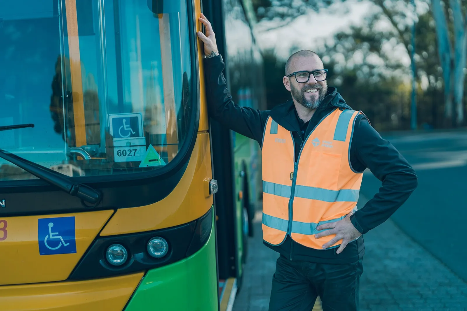 Smiling man in orange safety vest standing beside a green and yellow public bus with handicap signs.