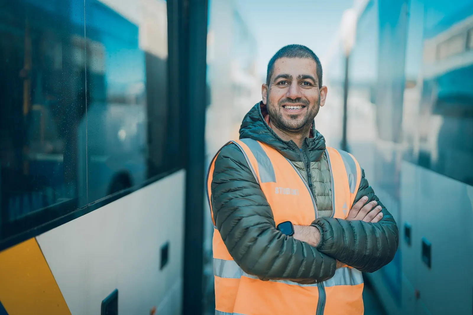 Smiling man in an orange safety vest with arms crossed standing between two white buses.