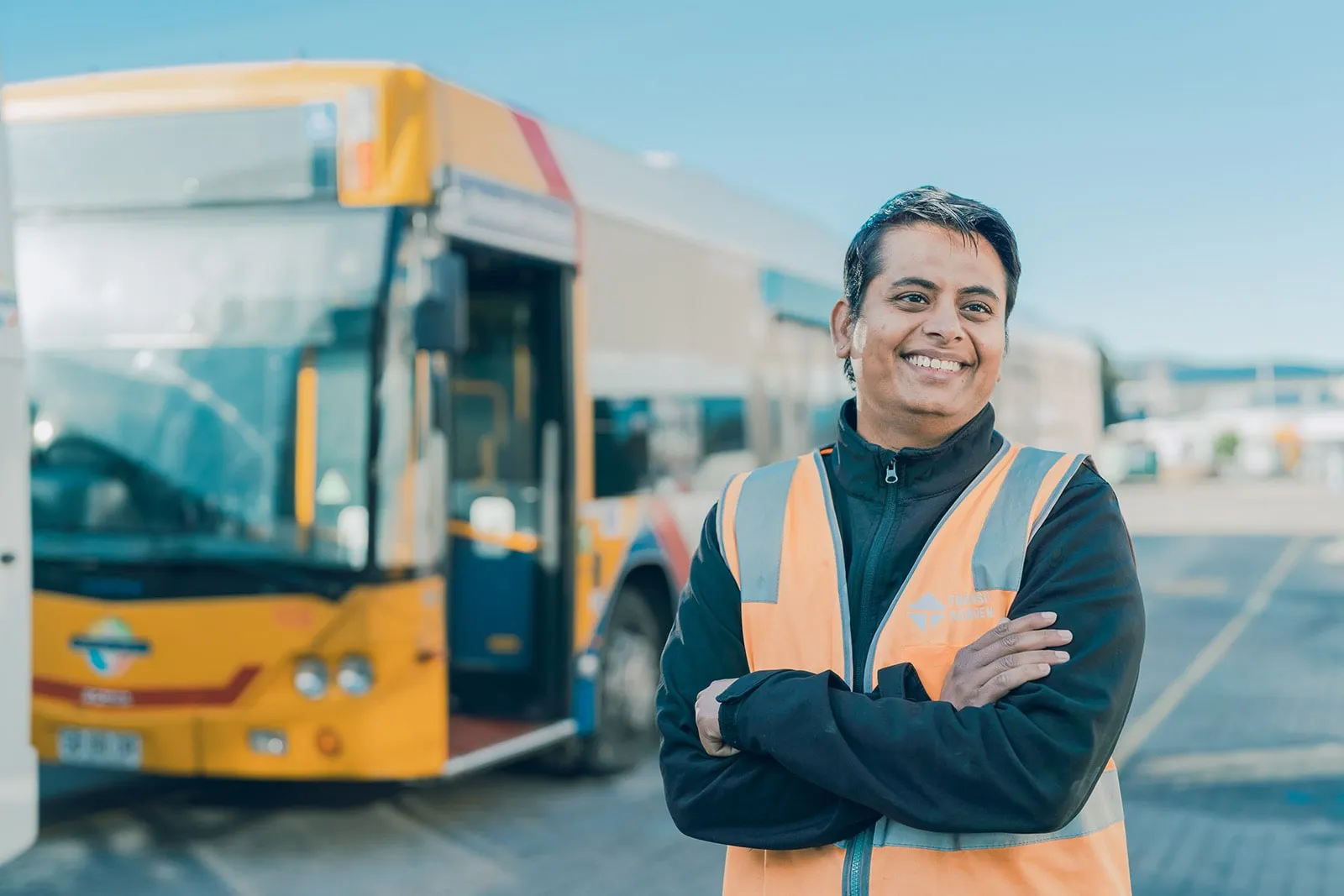 Smiling bus driver in orange safety vest standing with arms crossed in front of a yellow bus outdoors.