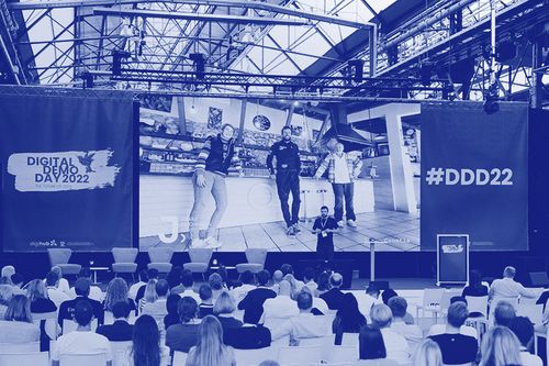 Audience seated in a large hall watching a presentation on a giant screen showing three people standing inside a modern industrial venue with banners reading Digital Demo Day 2022 and #DDD22.