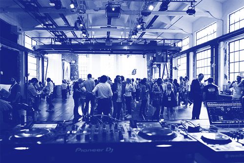 Crowd of people socializing in a spacious industrial-style venue with DJ equipment in the foreground.