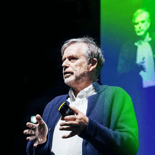 An older man with gray hair and beard wearing a white shirt and dark blue cardigan is speaking on stage with a microphone headset and holding a remote control.