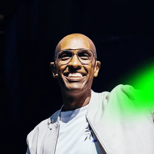 Smiling bald man with glasses wearing a white jacket and microphone headset against a dark background.