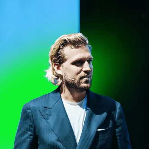 Blond man with beard wearing a dark blue suit jacket and white shirt against a blue to green gradient background.