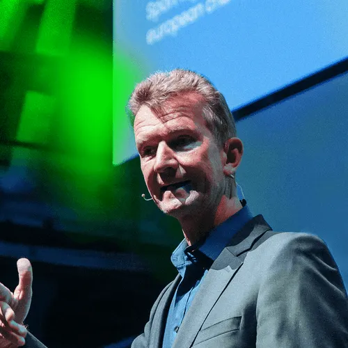 Man with short gray hair wearing a gray suit and blue shirt speaking on stage with a microphone headset.