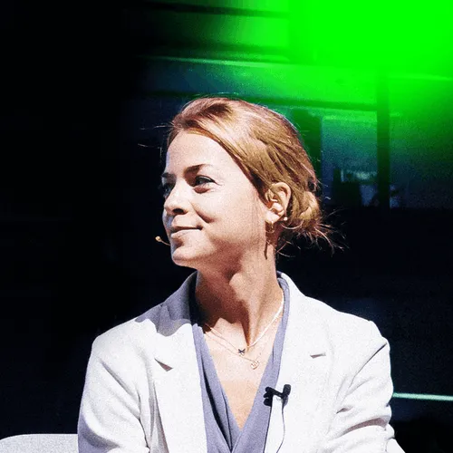 Woman with light brown hair in a bun wearing a white blazer and blue top, seated with a microphone headset, looking to the side.