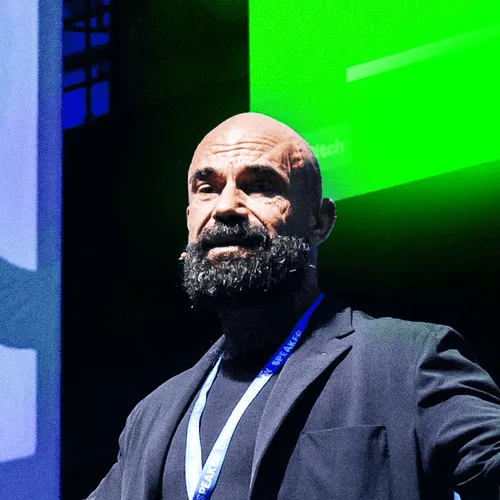 Bald man with a thick beard wearing a black blazer and a speaker lanyard on stage with green and blue background lighting.