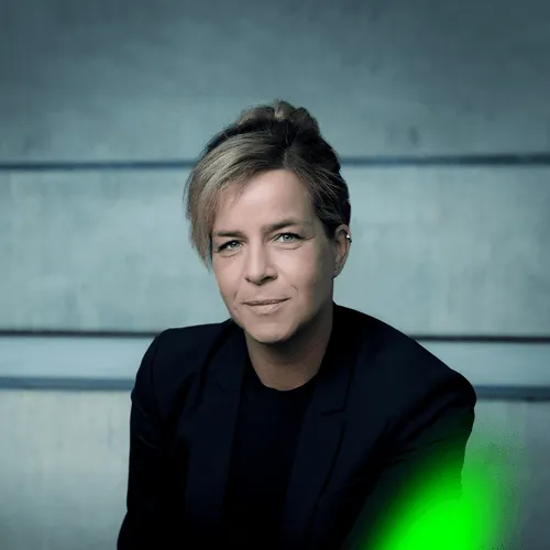 Portrait of a person with short blonde hair wearing a black blazer and sweater, sitting against a grey background.