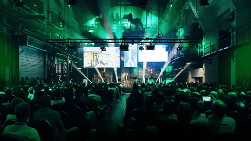 Large audience watching a speaker on stage with three screens and green lighting in an industrial-style venue.